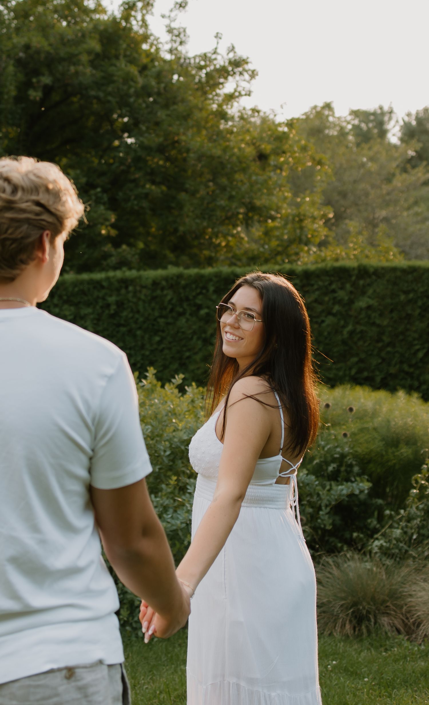 A smiling young woman in a white dress and glasses holds hands with a man in a garden at golden hour.