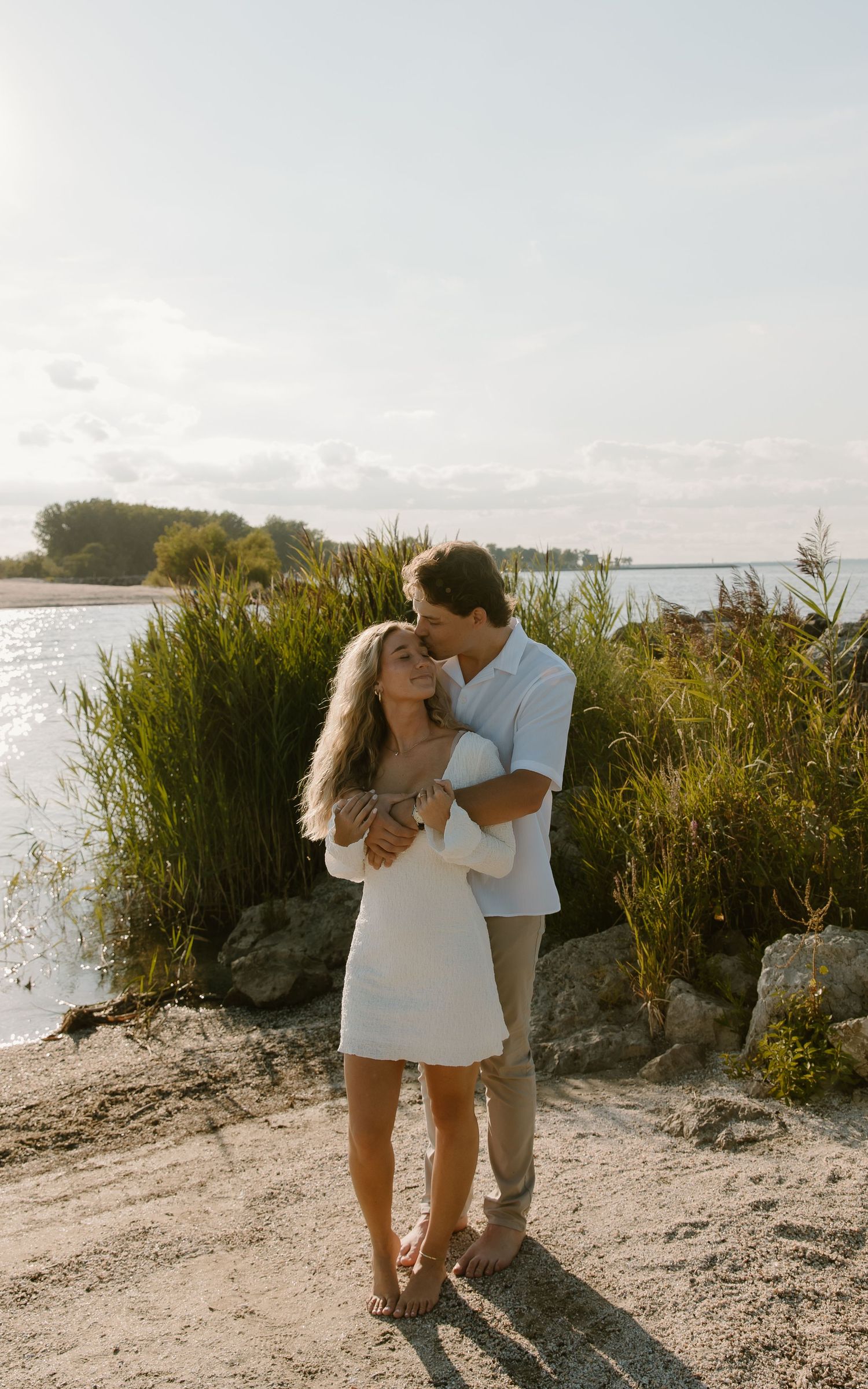 Couple embracing on sandy shore at golden hour, woman in white dress, lush greenery and water in background.