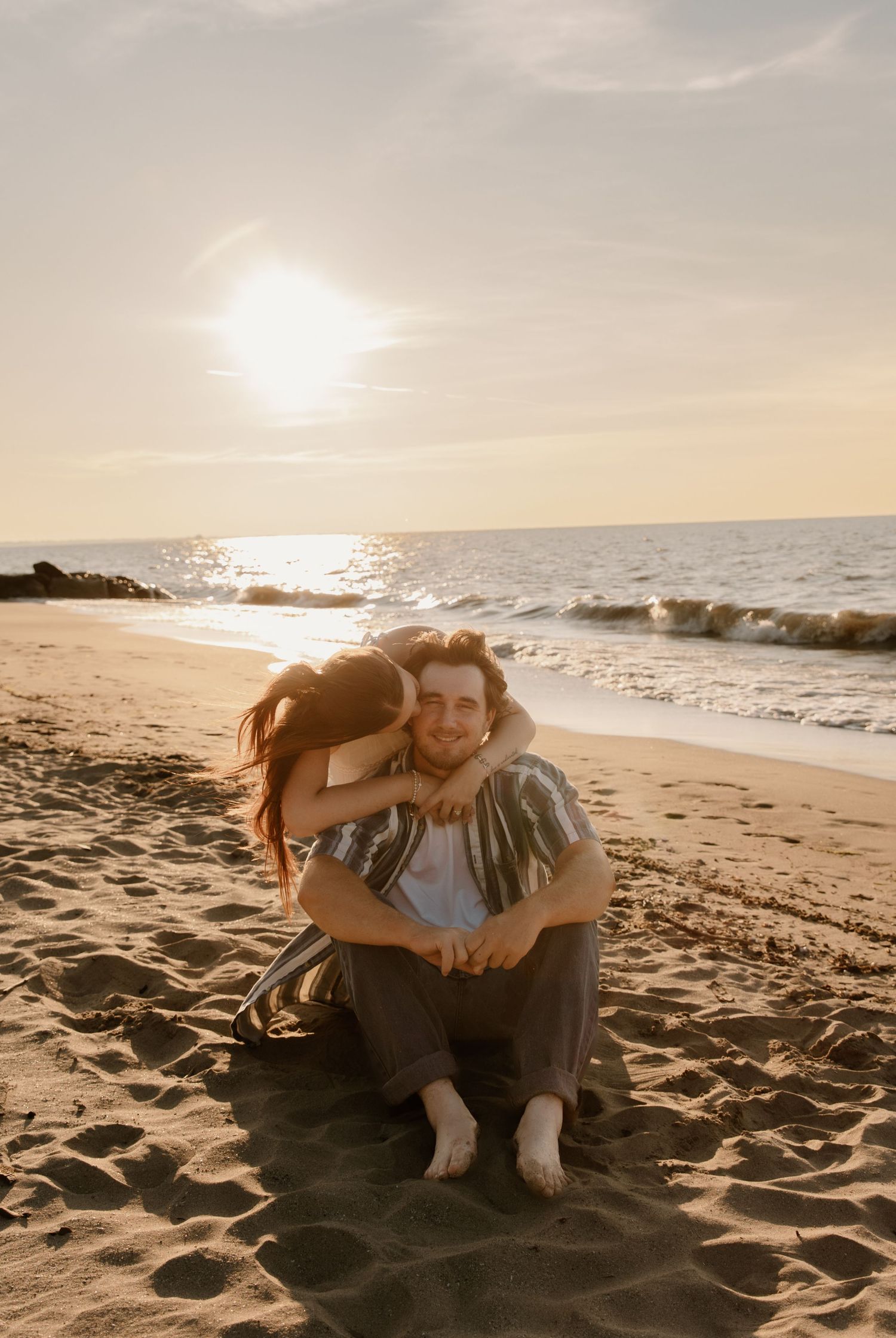 Couple sitting on sandy beach at sunset, woman hugging man from behind, ocean waves in background.