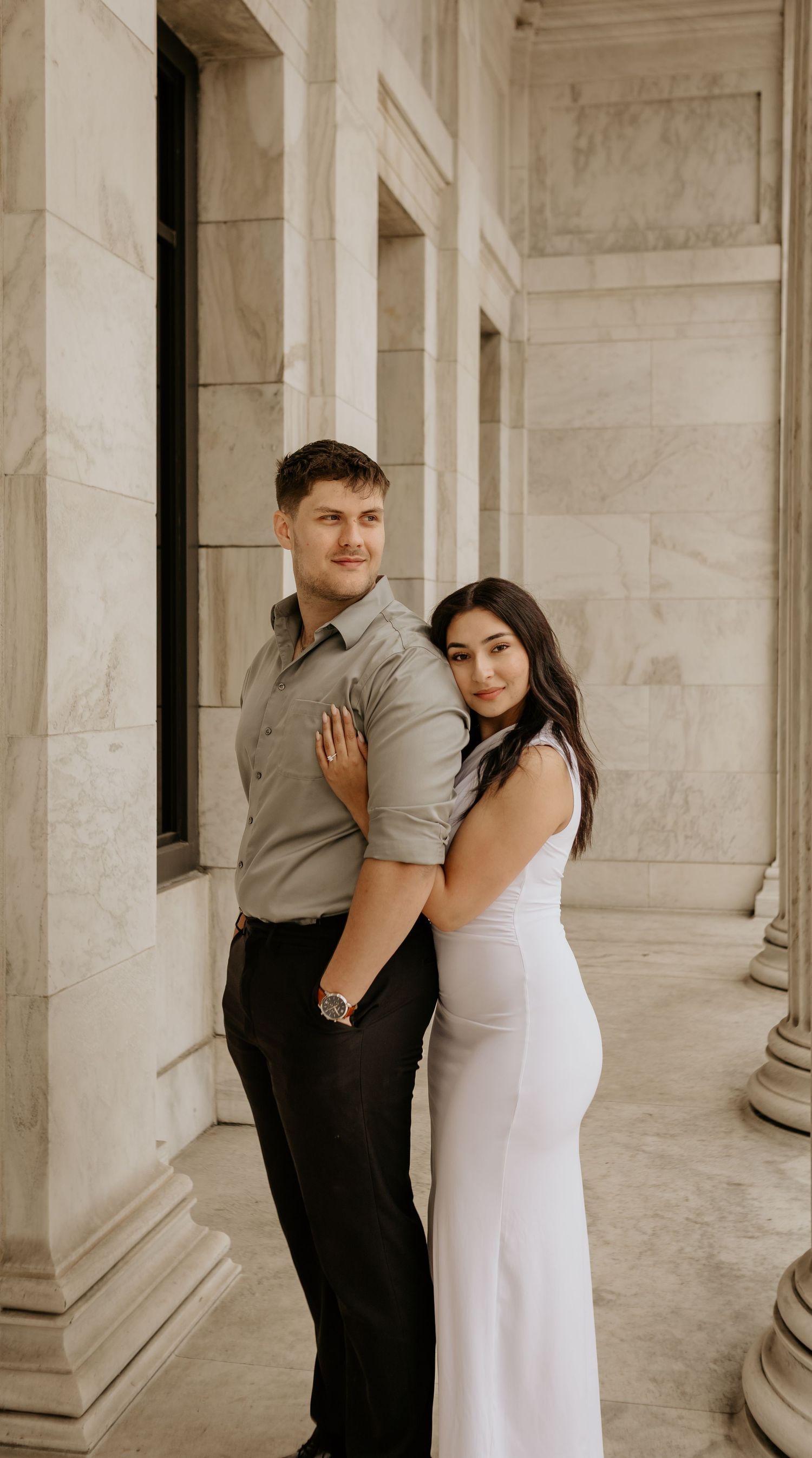 A couple in formal attire poses together against marble columns.