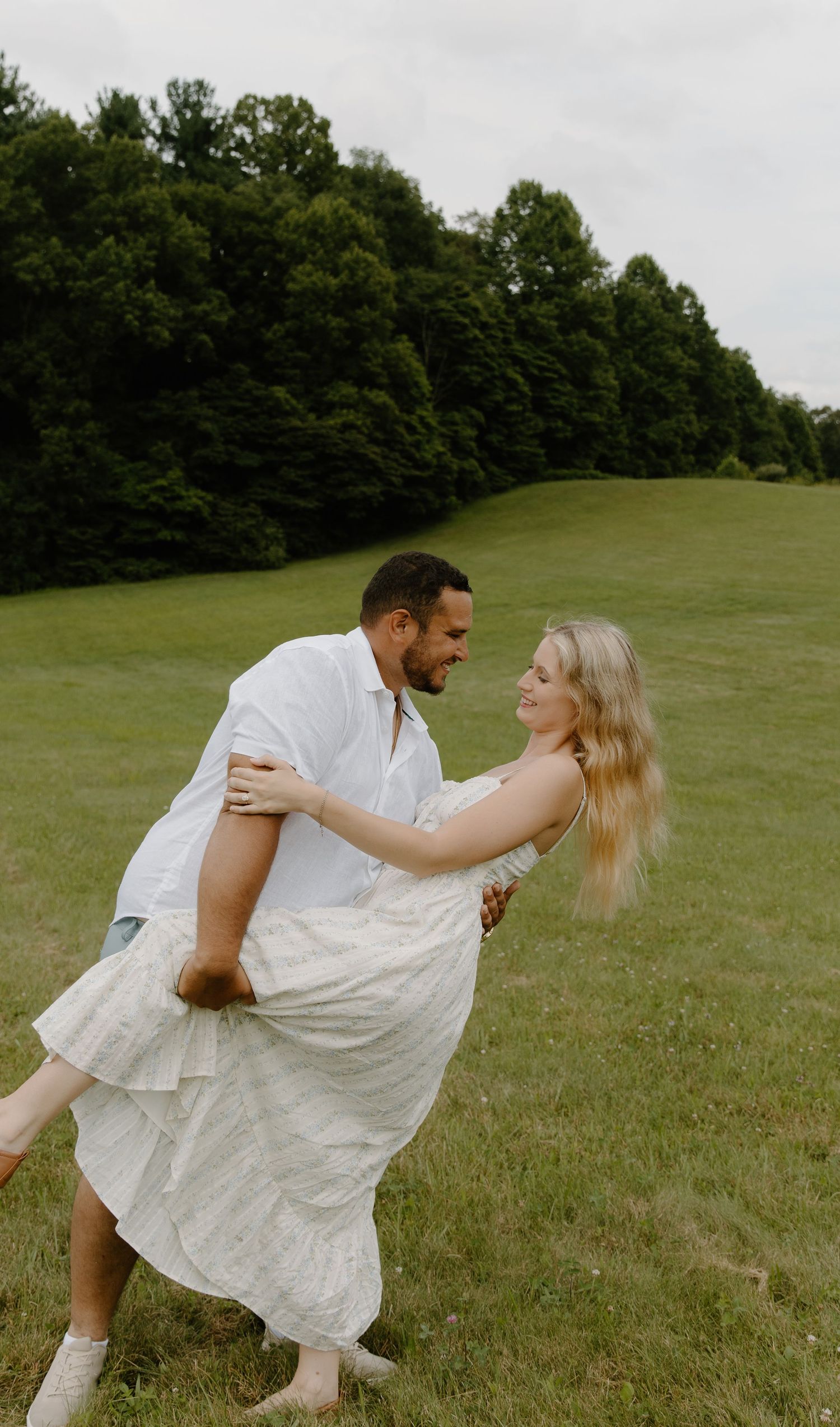 A couple dances together in a grassy field on a cloudy summer day, with trees in the cuyahoga falls background.