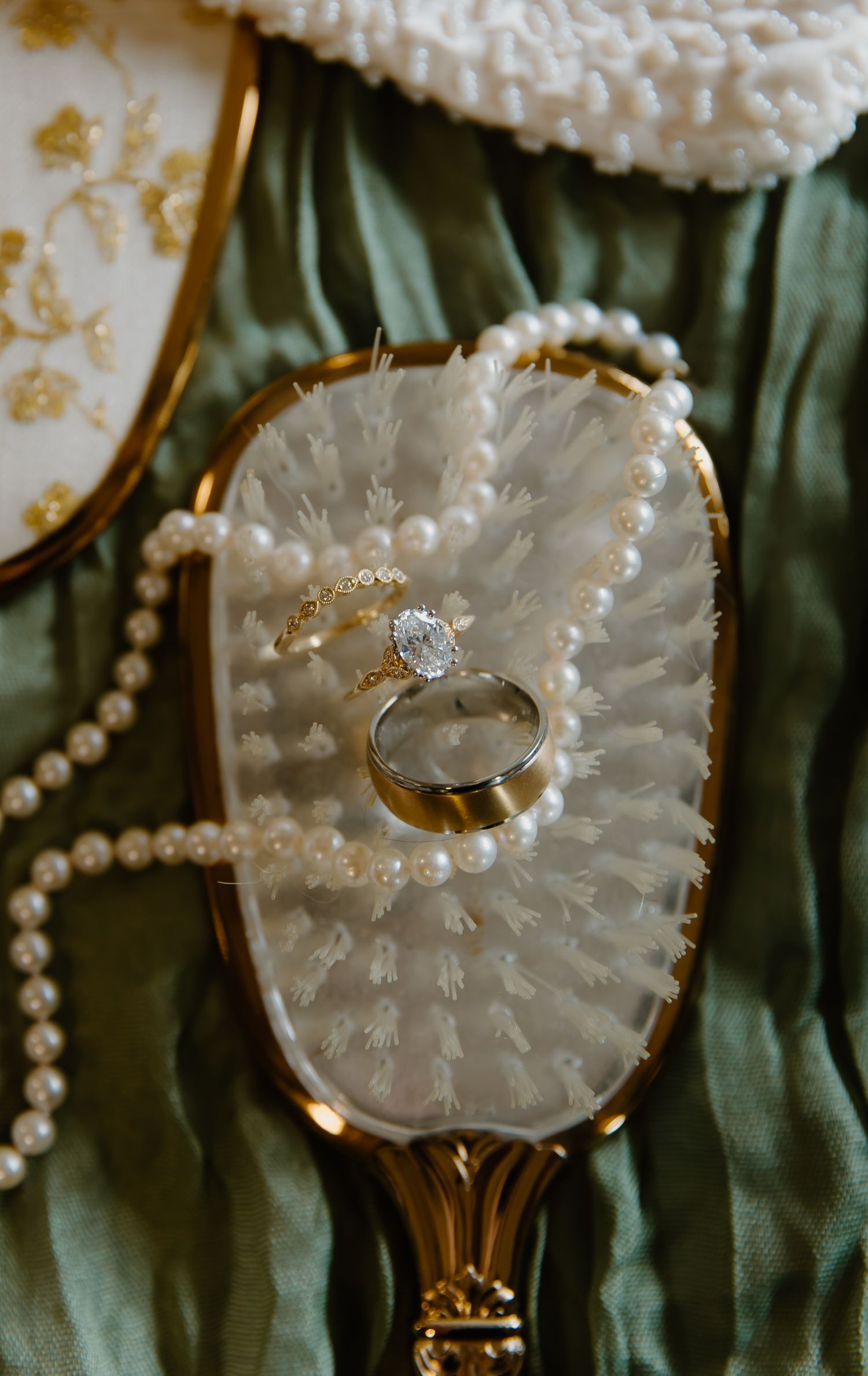 Gold engagement ring with oval diamond and wedding band on vintage mirror with pearls and white feathers.
