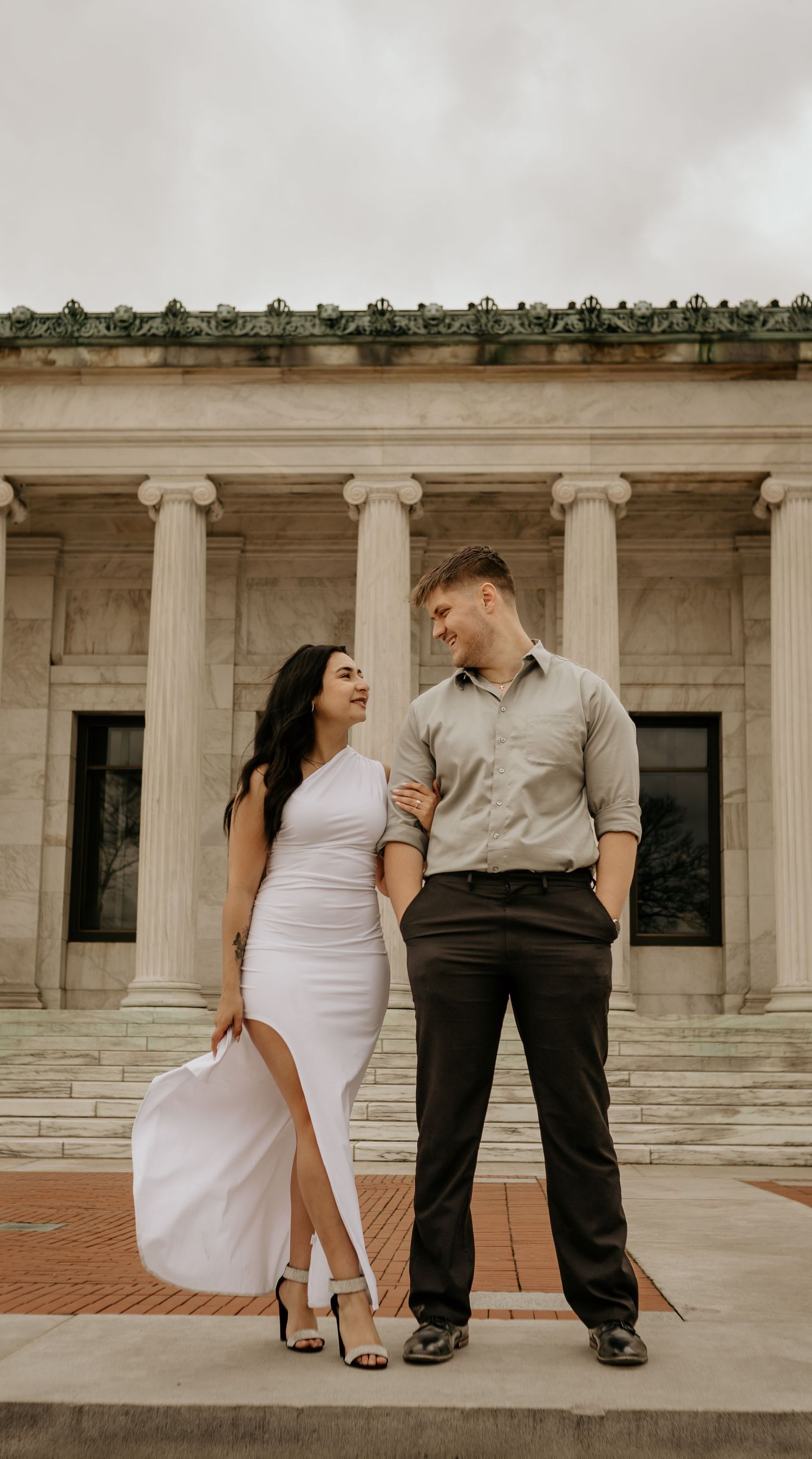 A couple stands together in front of classical Greek columns and architecture.