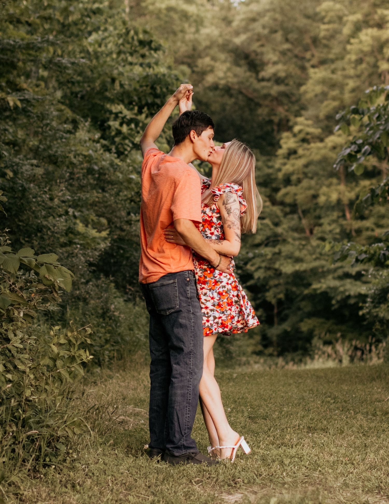 A couple embraces romantically in a garden setting with her wearing a floral dress and him in an orange shirt.