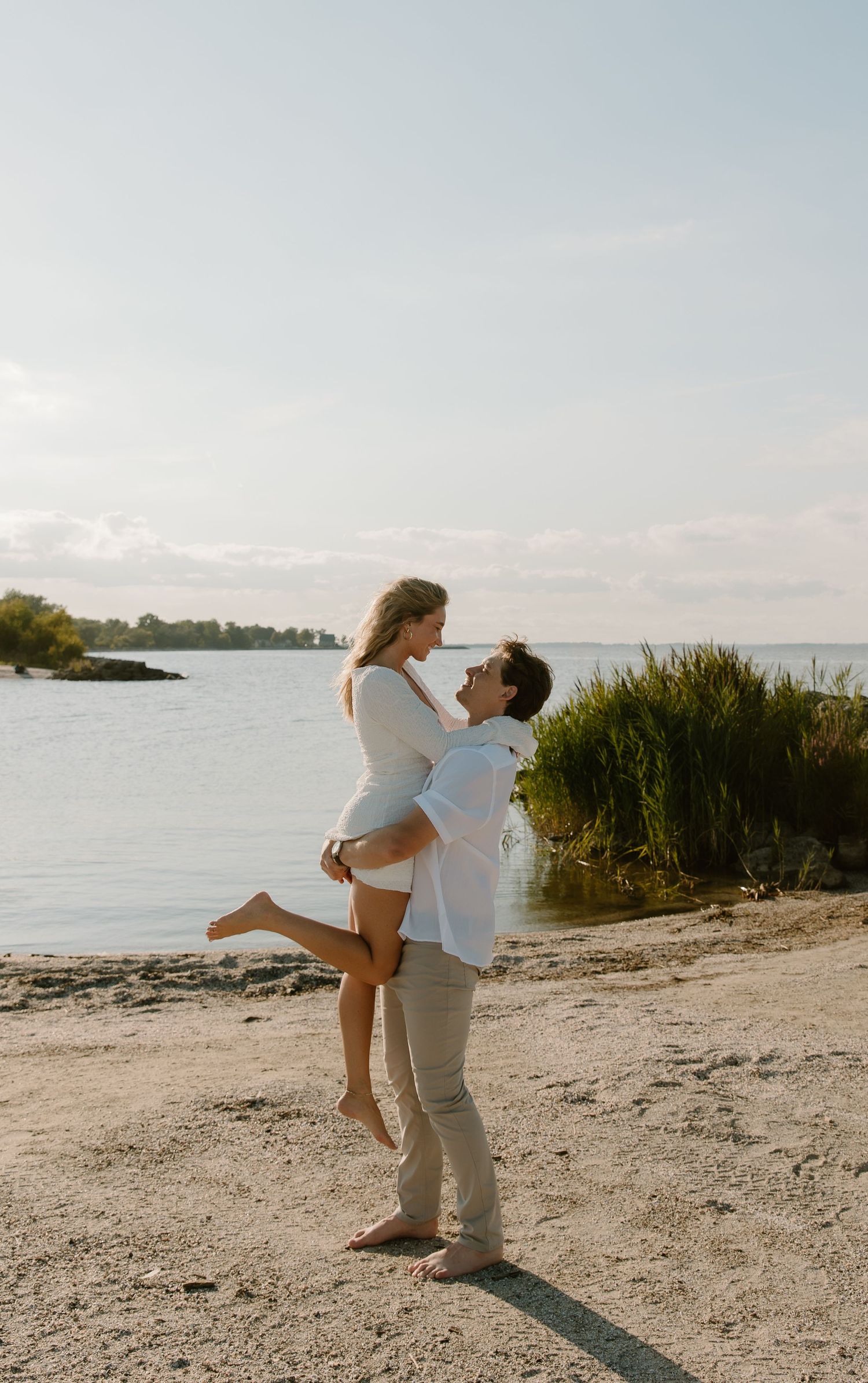 A man lifts a woman on a sandy beach at sunset, both smiling in white outfits near calm water.