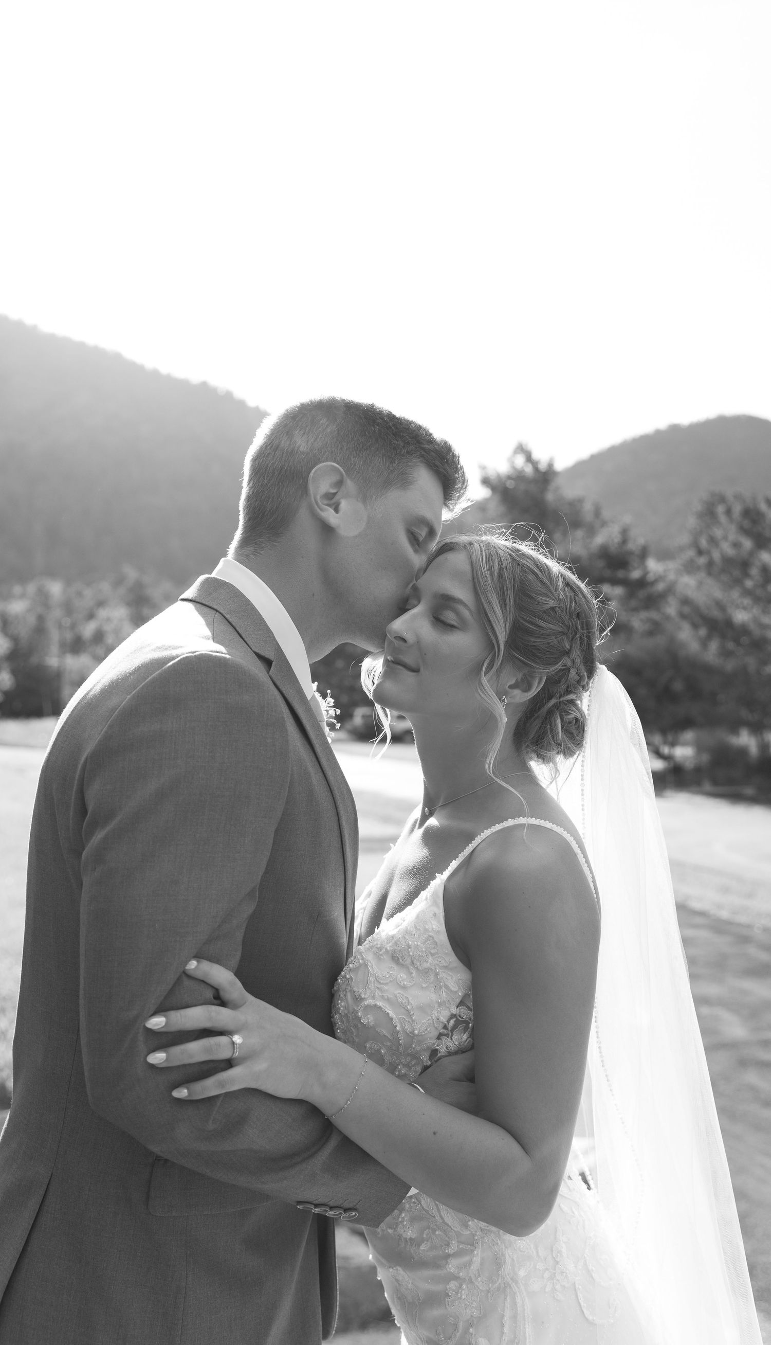 A romantic black and white wedding photo showing a tender moment between newlyweds against a mountain backdrop.