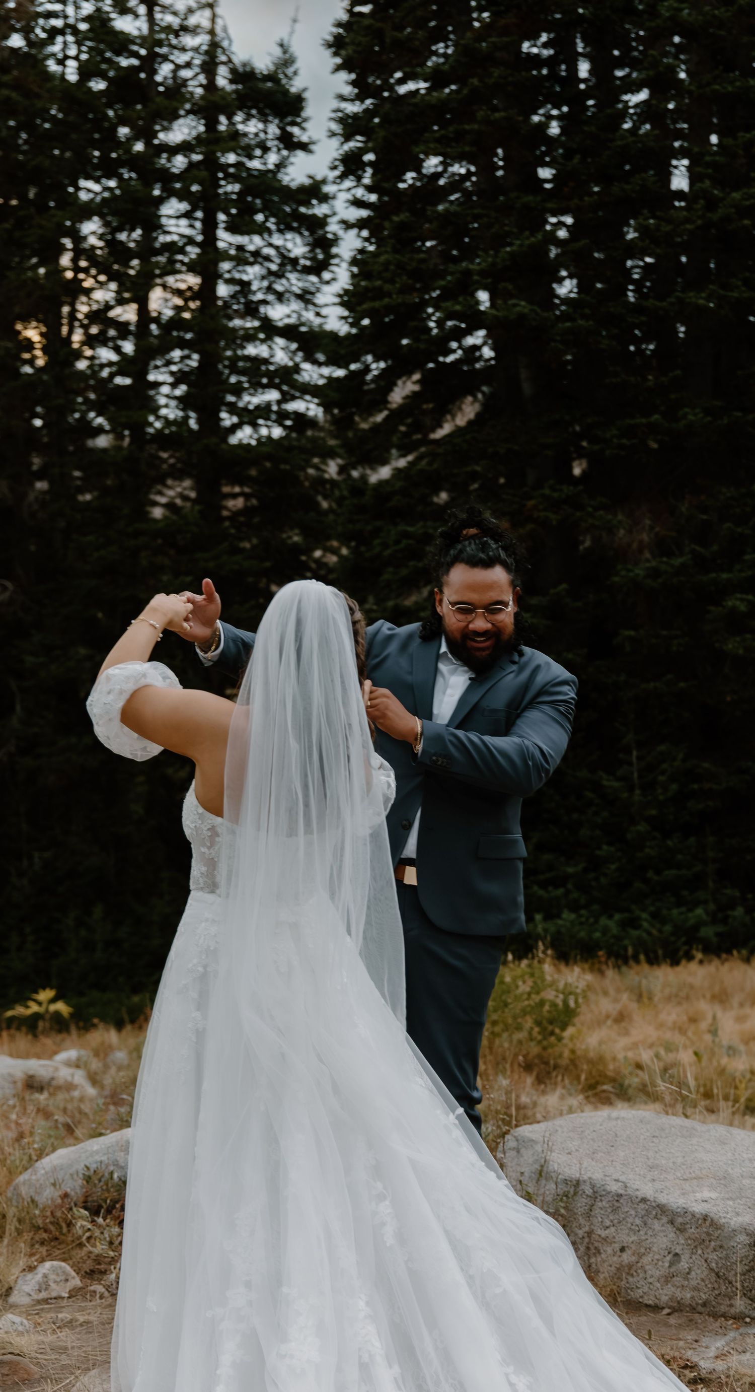 A couple in wedding attire stands in front of evergreen trees while adjusting the bride's flowing veil.