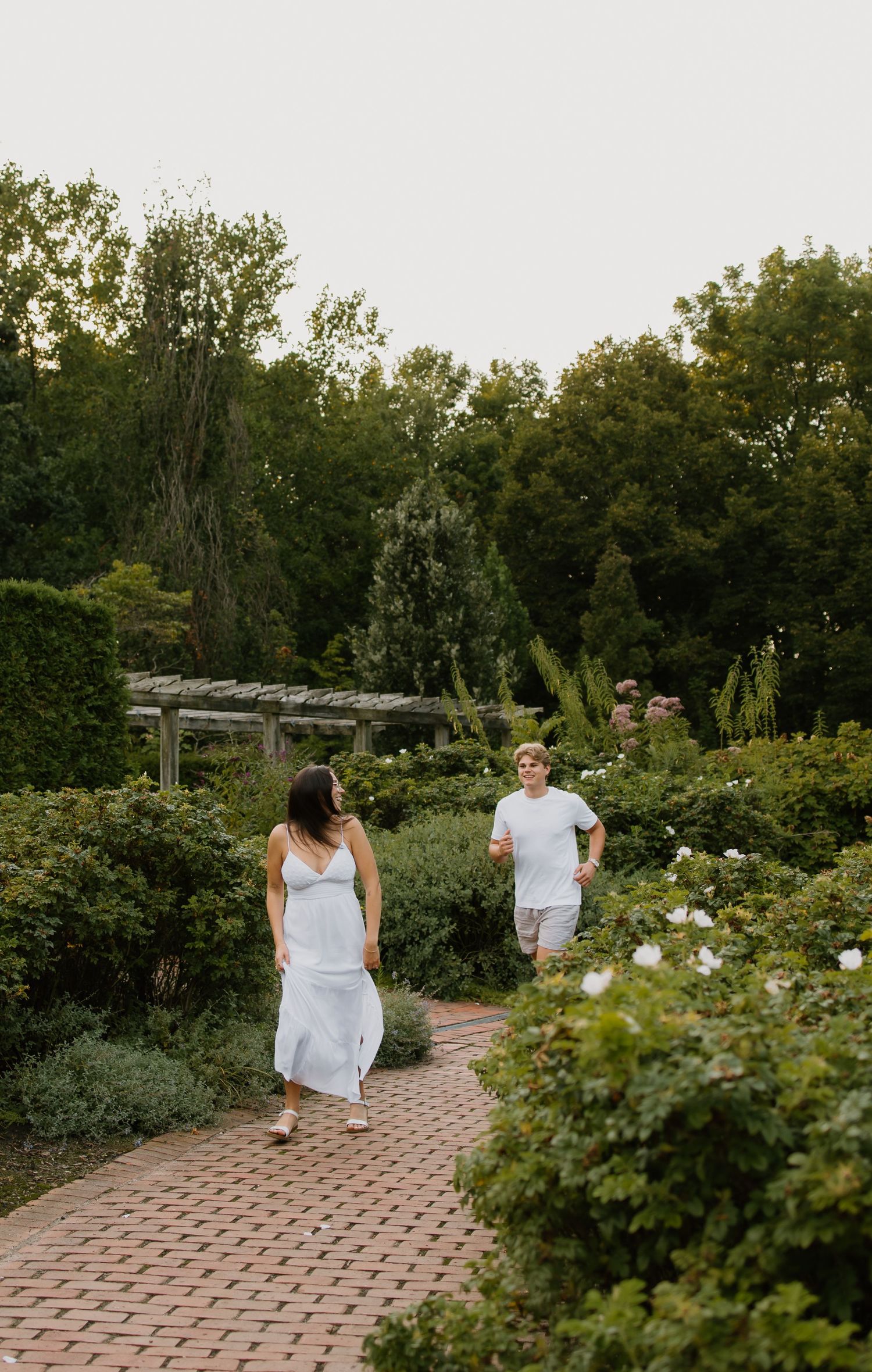 A couple in white outfits strolls along a brick garden path surrounded by lush greenery and blooming flowers.