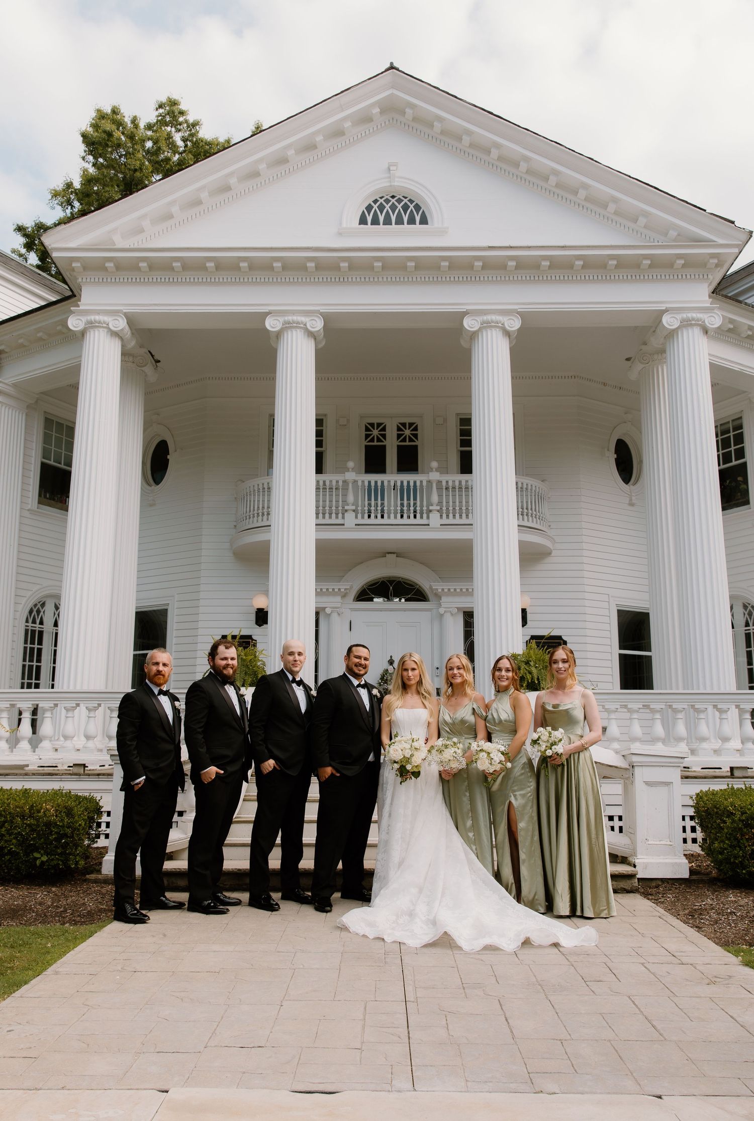 Wedding party poses in front of elegant white colonial venue, bridesmaids in sage green gowns.