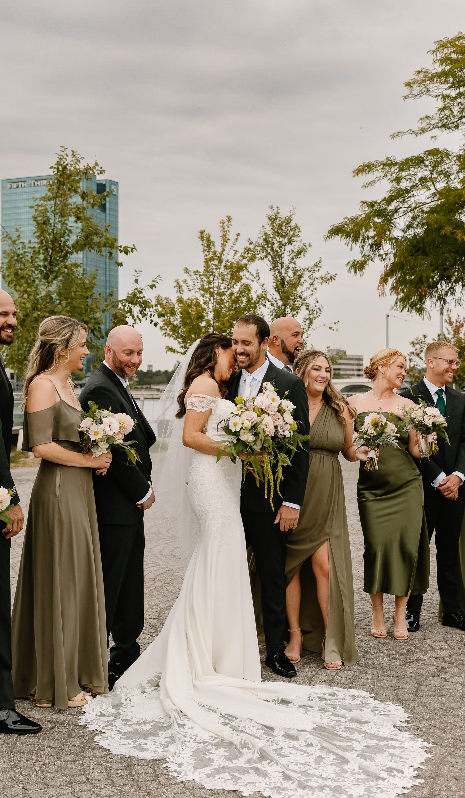 Wedding party celebrates on outdoor terrace with bridesmaids in olive dresses and groomsmen in black suits.