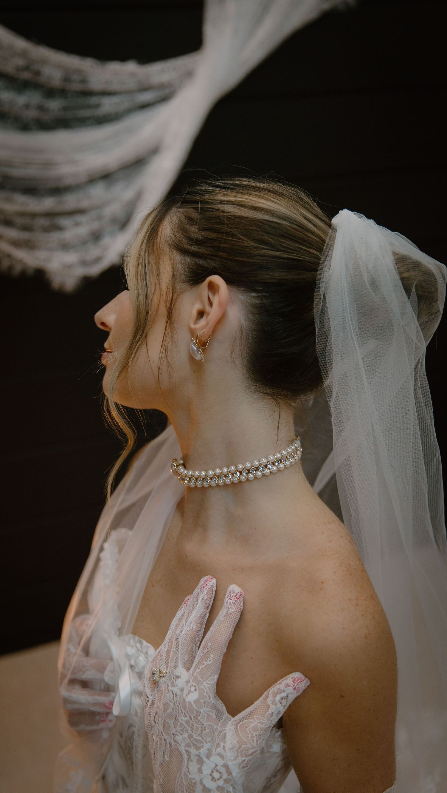 Close-up details of a bride wearing a pearl necklace, lace wedding dress and flowing veil against dark background.