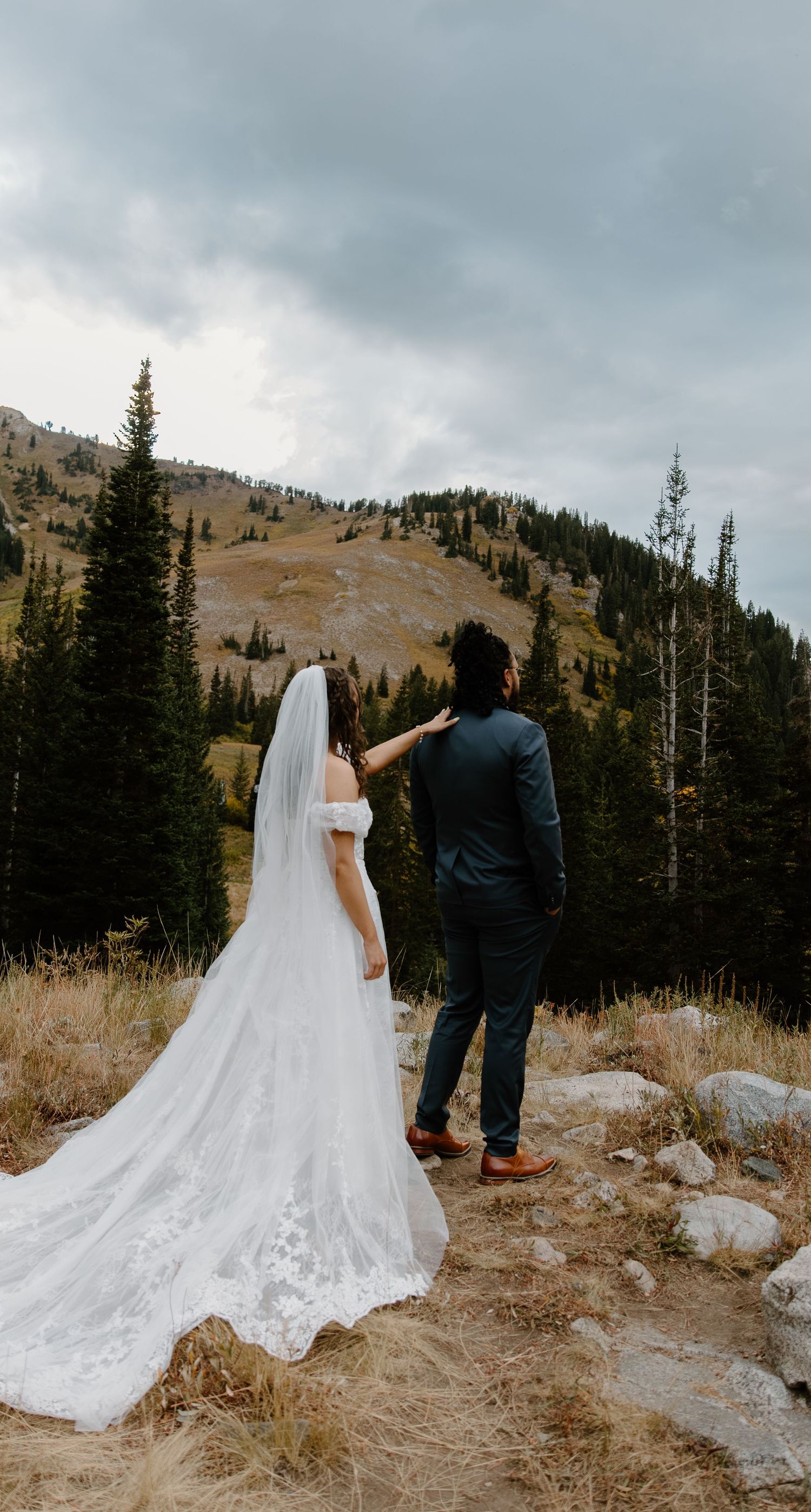 A couple in wedding attire stands on a mountain trail overlooking a scenic valley with evergreen trees and cloudy skies.