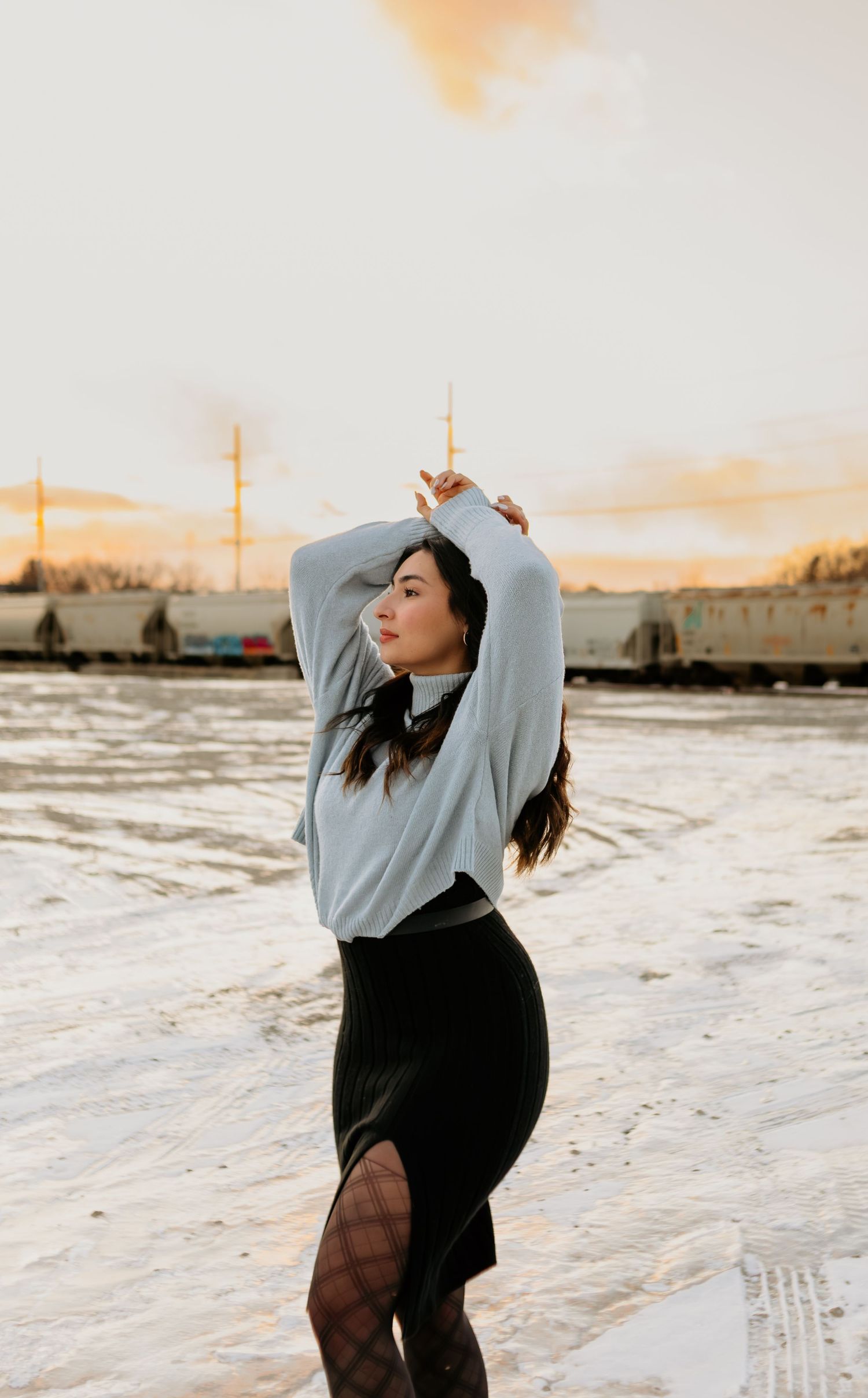 Person in grey sweater poses dramatically at sunset with train cars in background.