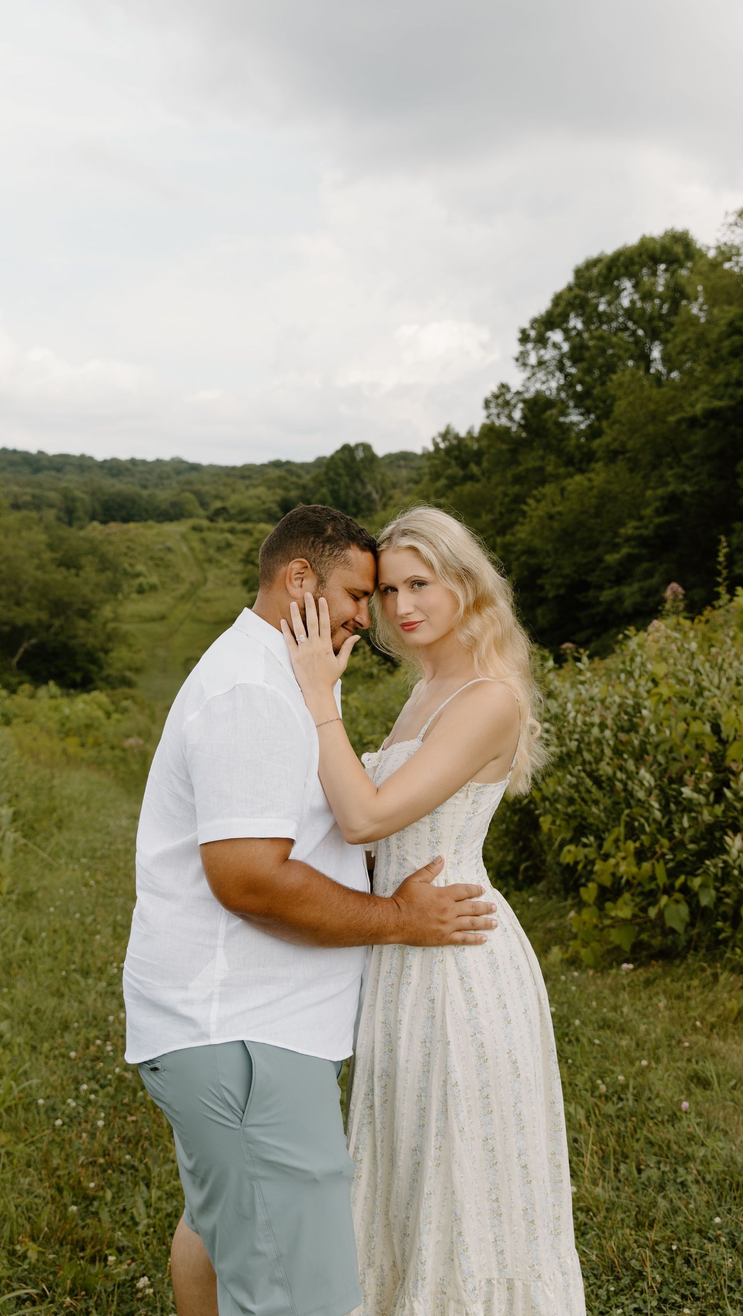 A couple shares an intimate moment in a scenic meadow, dressed in white against a backdrop of lush green cuyahoga falls hills.