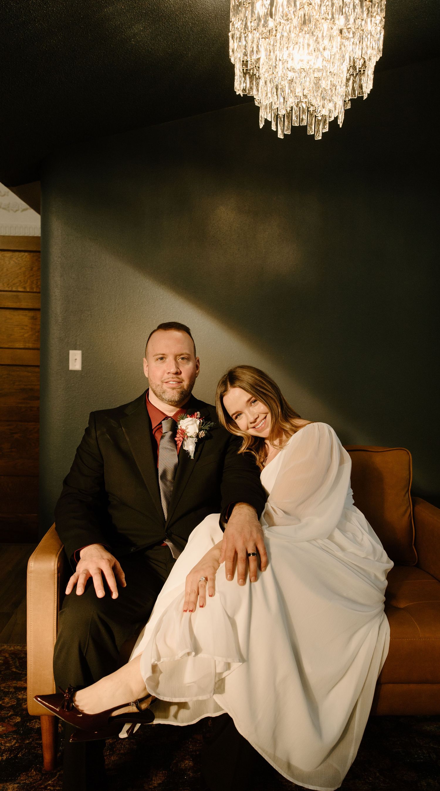 A romantic couple poses together on a couch under elegant crystal chandeliers in a dimly lit room.