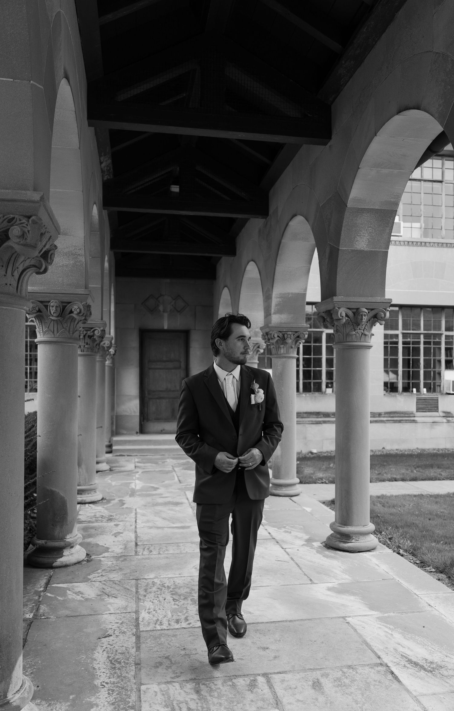 Groom in a dark suit walks under ornate stone arches in a black and white wedding photo.