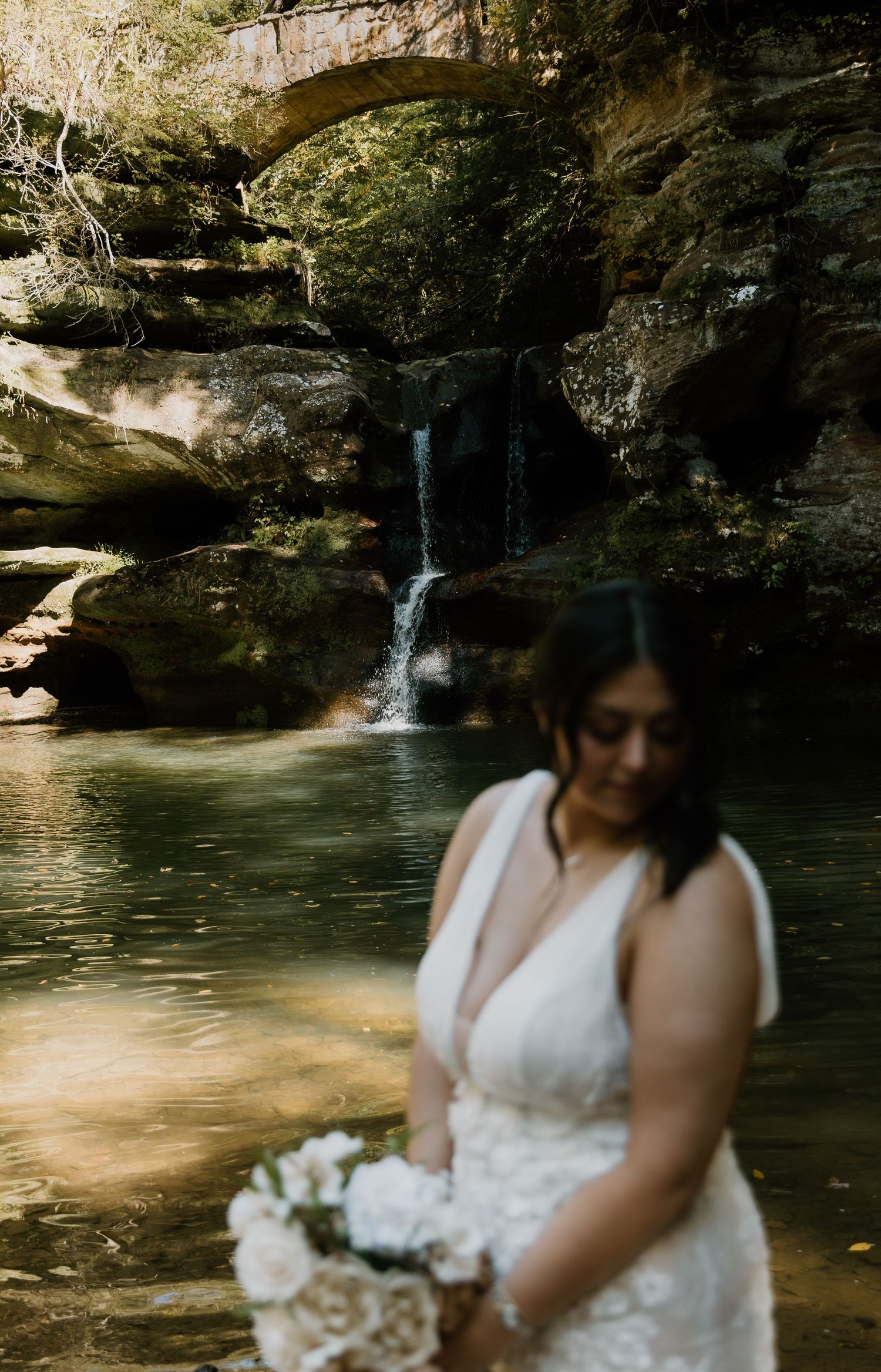 Bride in lace gown holds white bouquet near waterfall and stone bridge in Hocking Hills, Ohio.