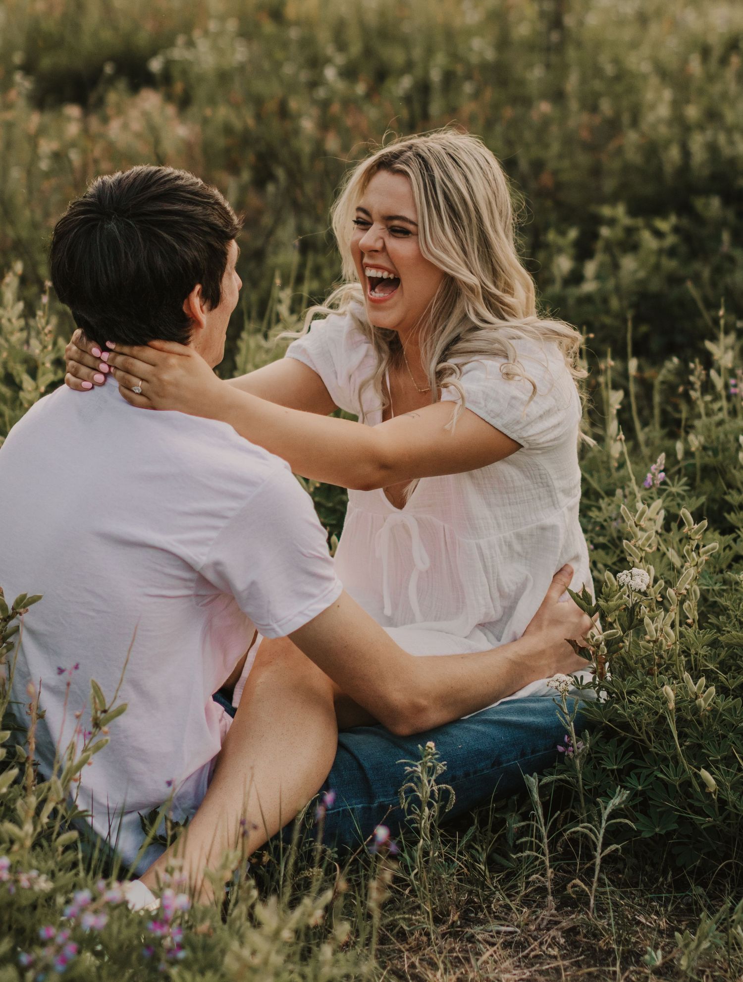 A joyful couple laughs together while sitting in a wildflower meadow at sunset.