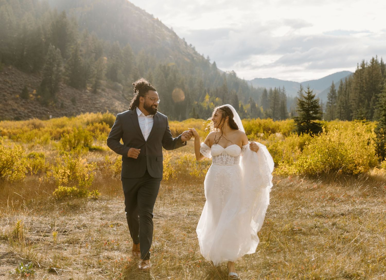 A couple in wedding attire walks and dances through a scenic mountain meadow with golden fall foliage at sunset.