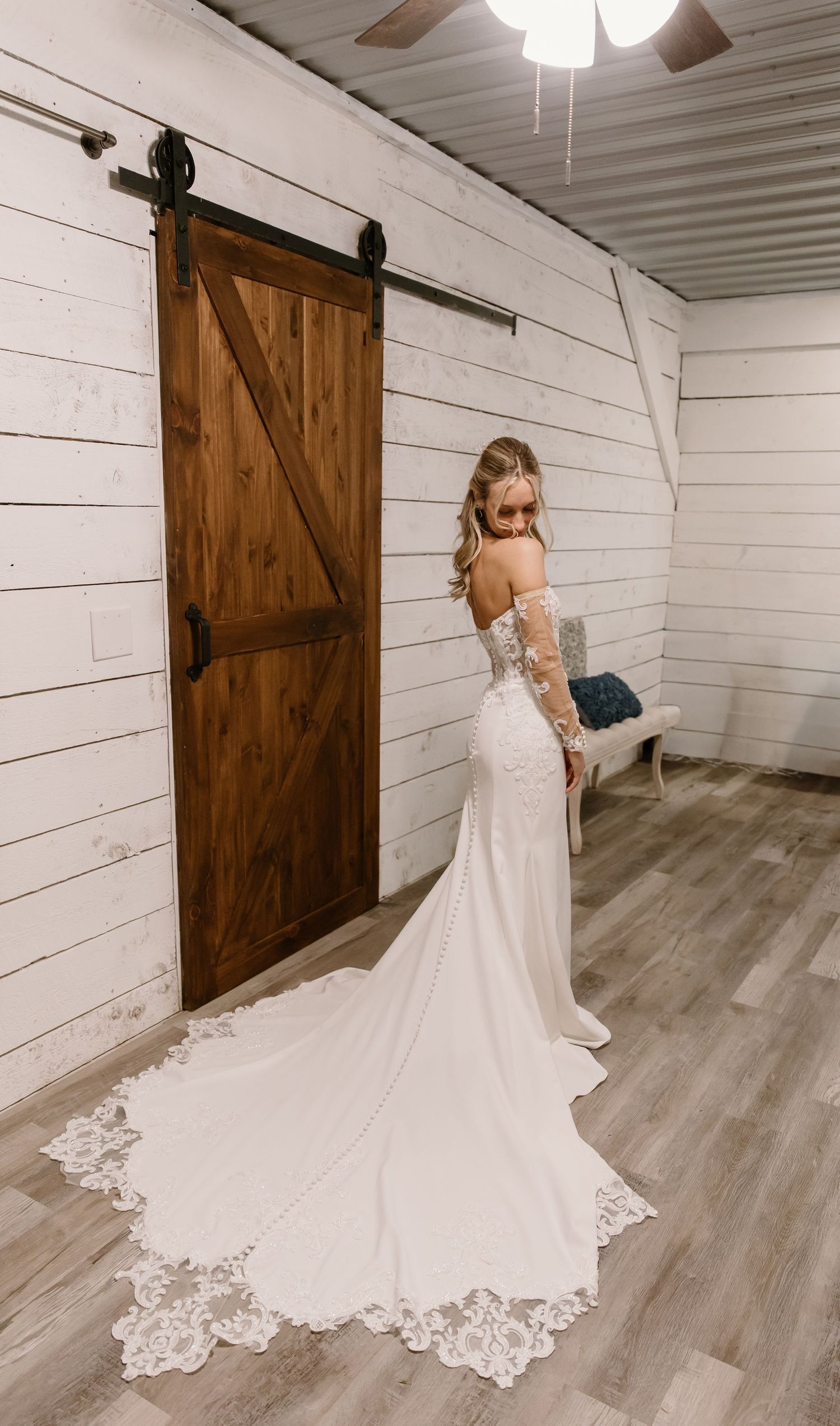 Bride in elegant lace wedding gown with long train poses by rustic barn door on wood floors.