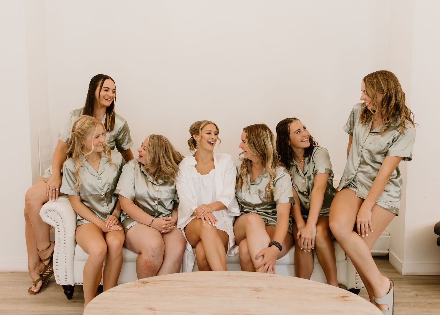 A group of women in casual attire sitting together on a wooden floor against a white wall.