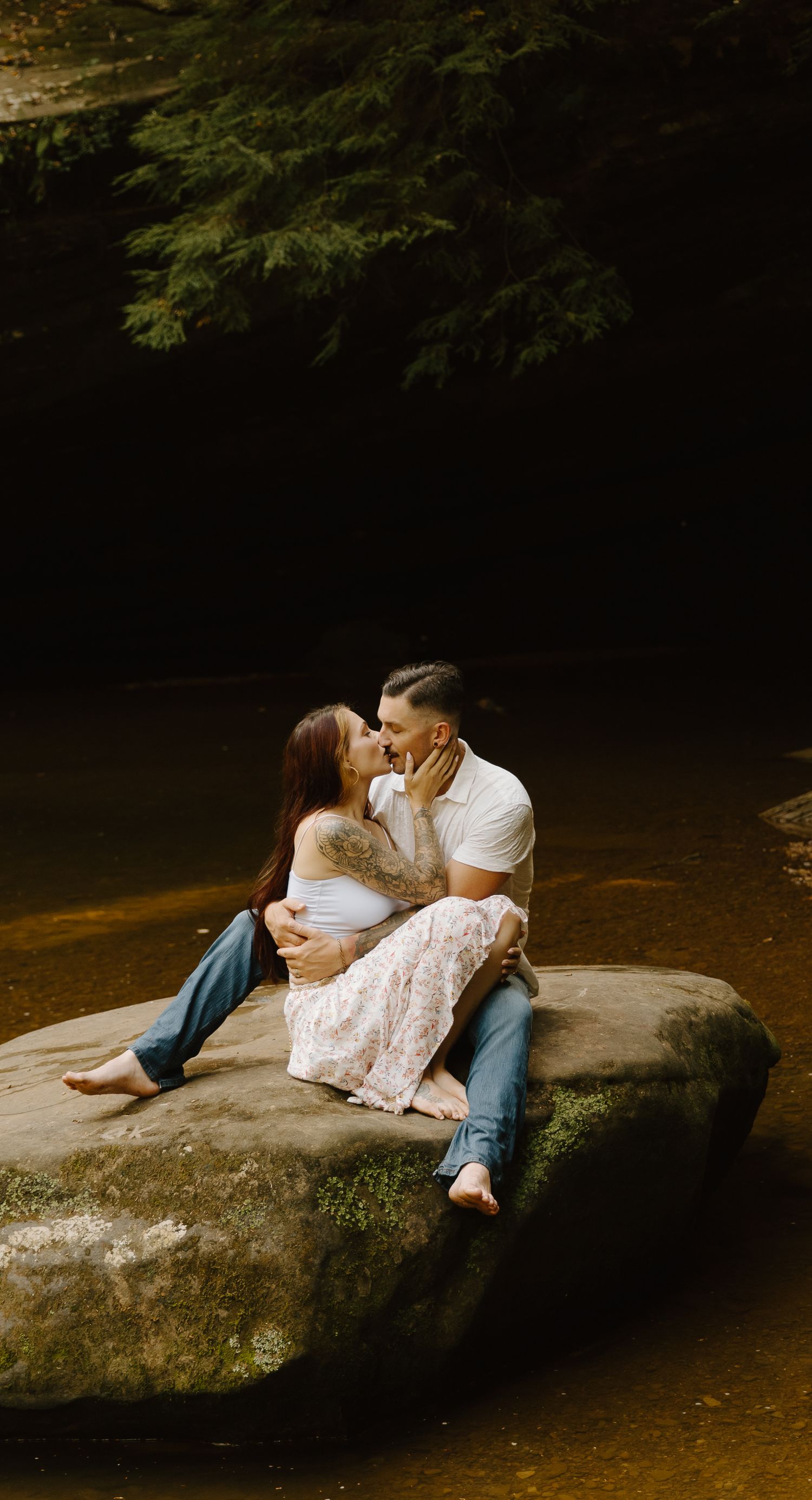 Couple sharing an intimate moment while sitting on a large rock by a serene river in a forest setting.