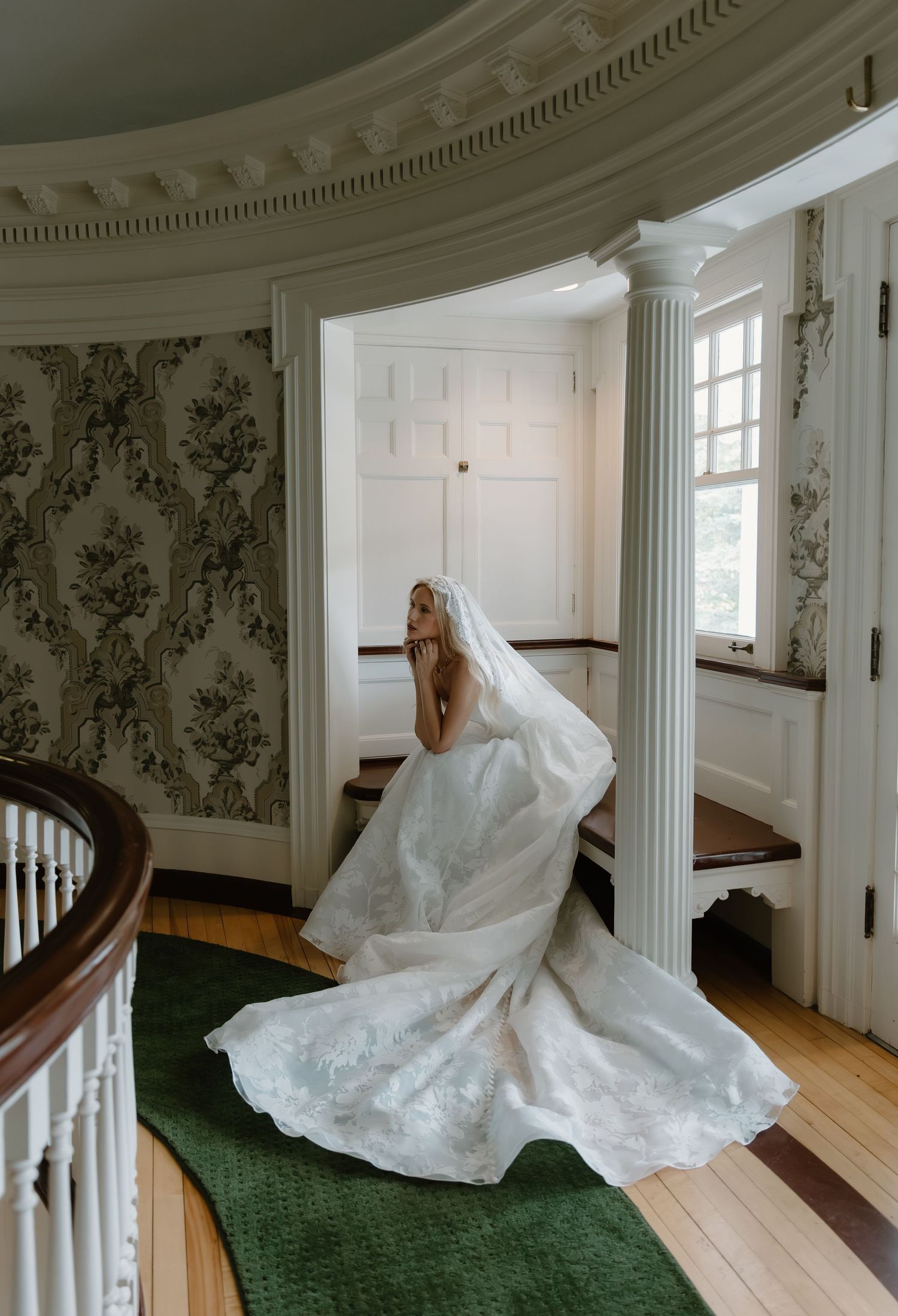 Bride in a flowing lace wedding gown and veil standing in a grand historic mansion foyer.