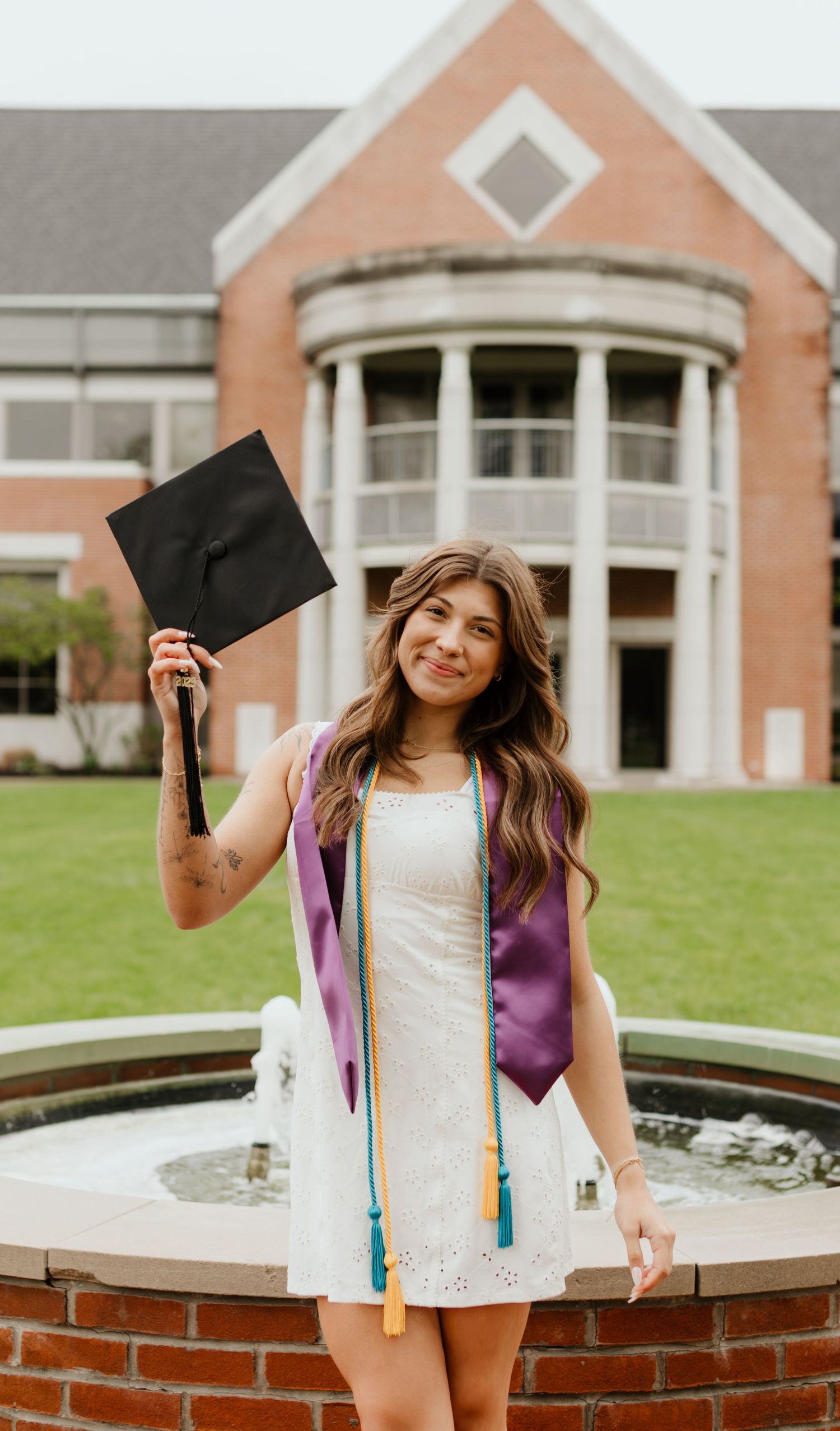 A graduate in a white dress and purple graduation stole holds up their cap in front of a brick building with a fountain.