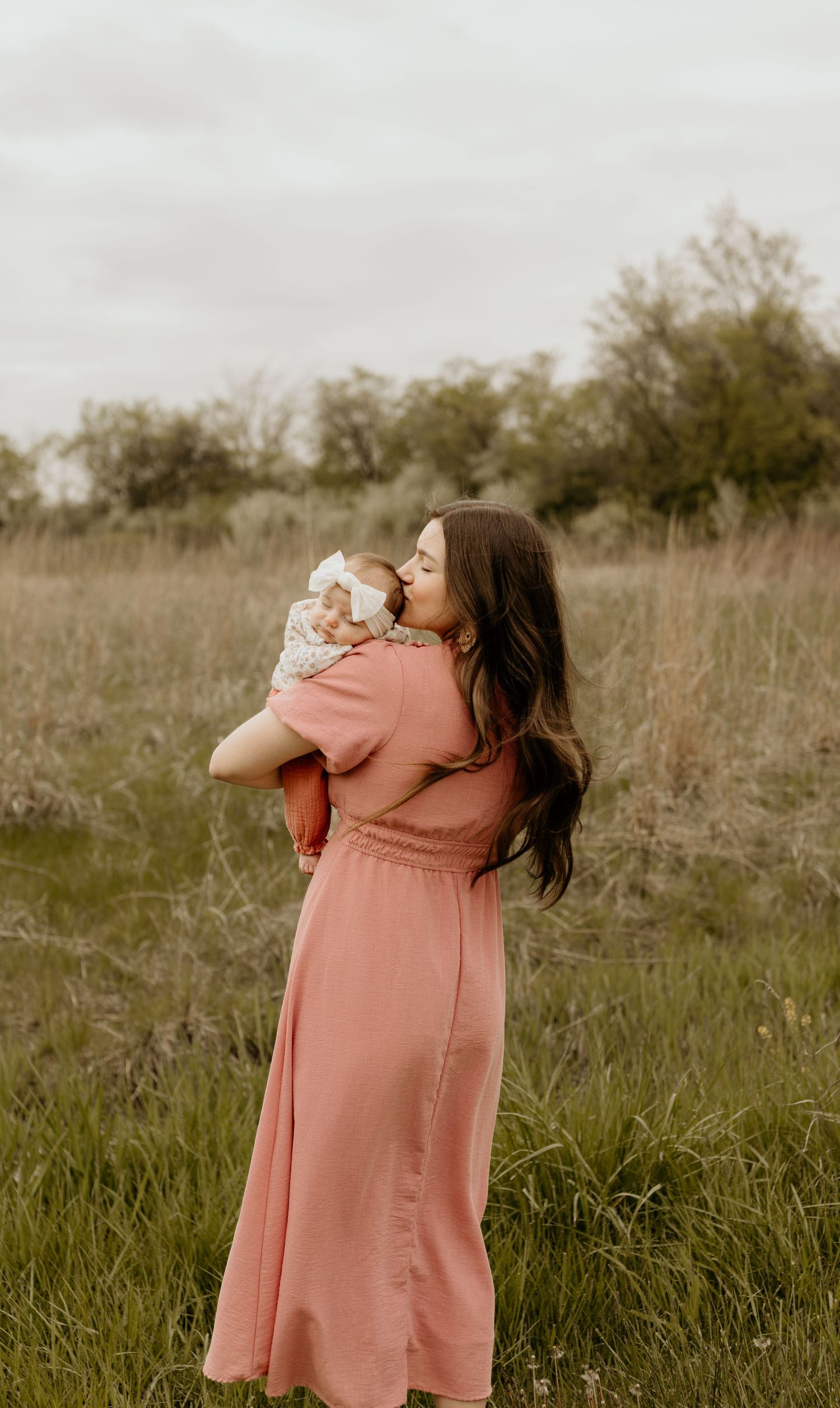 Person in a peach-colored maxi dress stands in a sunlit meadow with tall grass and trees in the background.
