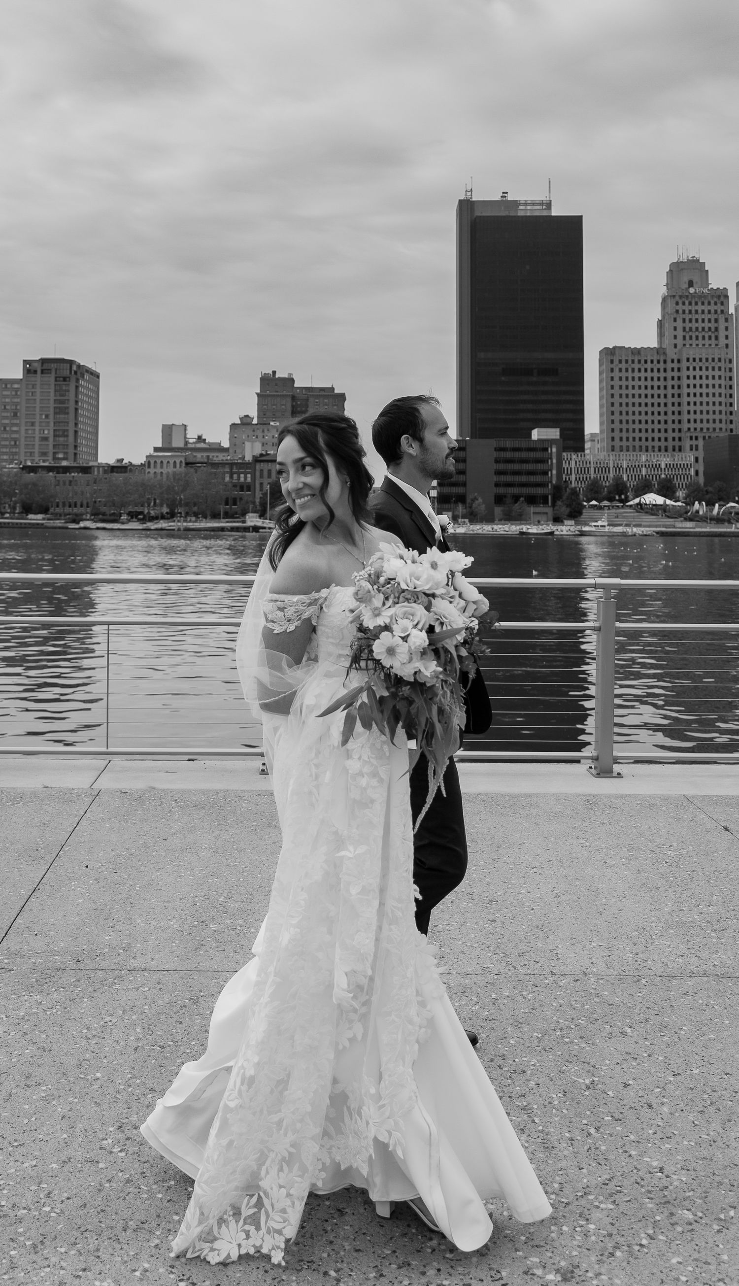 Elegant wedding couple dances on waterfront plaza with urban skyline backdrop in black and white.