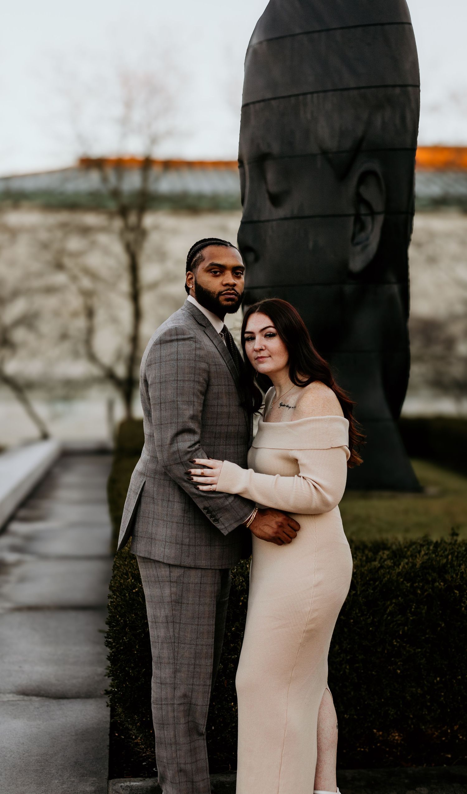 Stylish couple embracing outside near modern sculpture on rainy day.