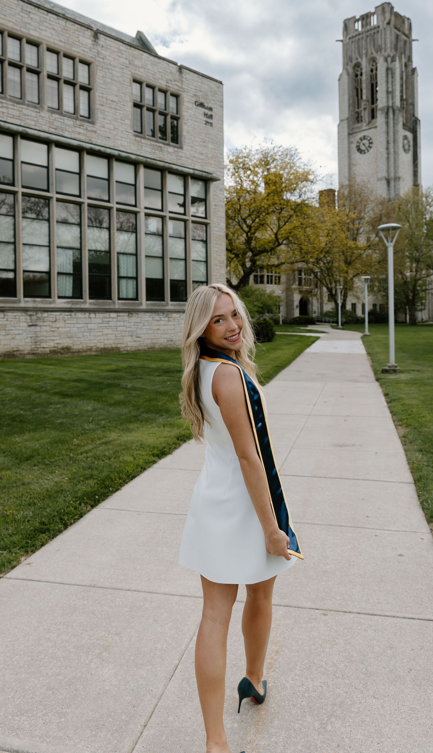 A smiling blonde woman in a white dress holds a graduation stole on a college campus sidewalk with a clock tower behind her.