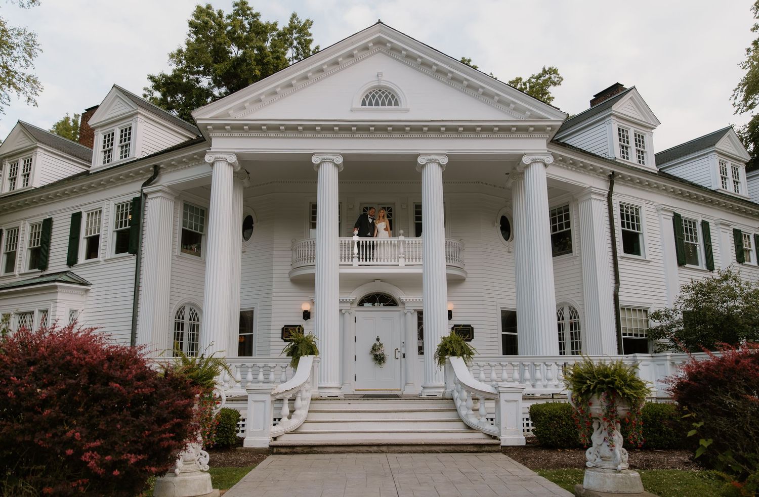 Elegant white neoclassical mansion with grand columns, balcony, and symmetrical staircase entrance surrounded by lush greenery.