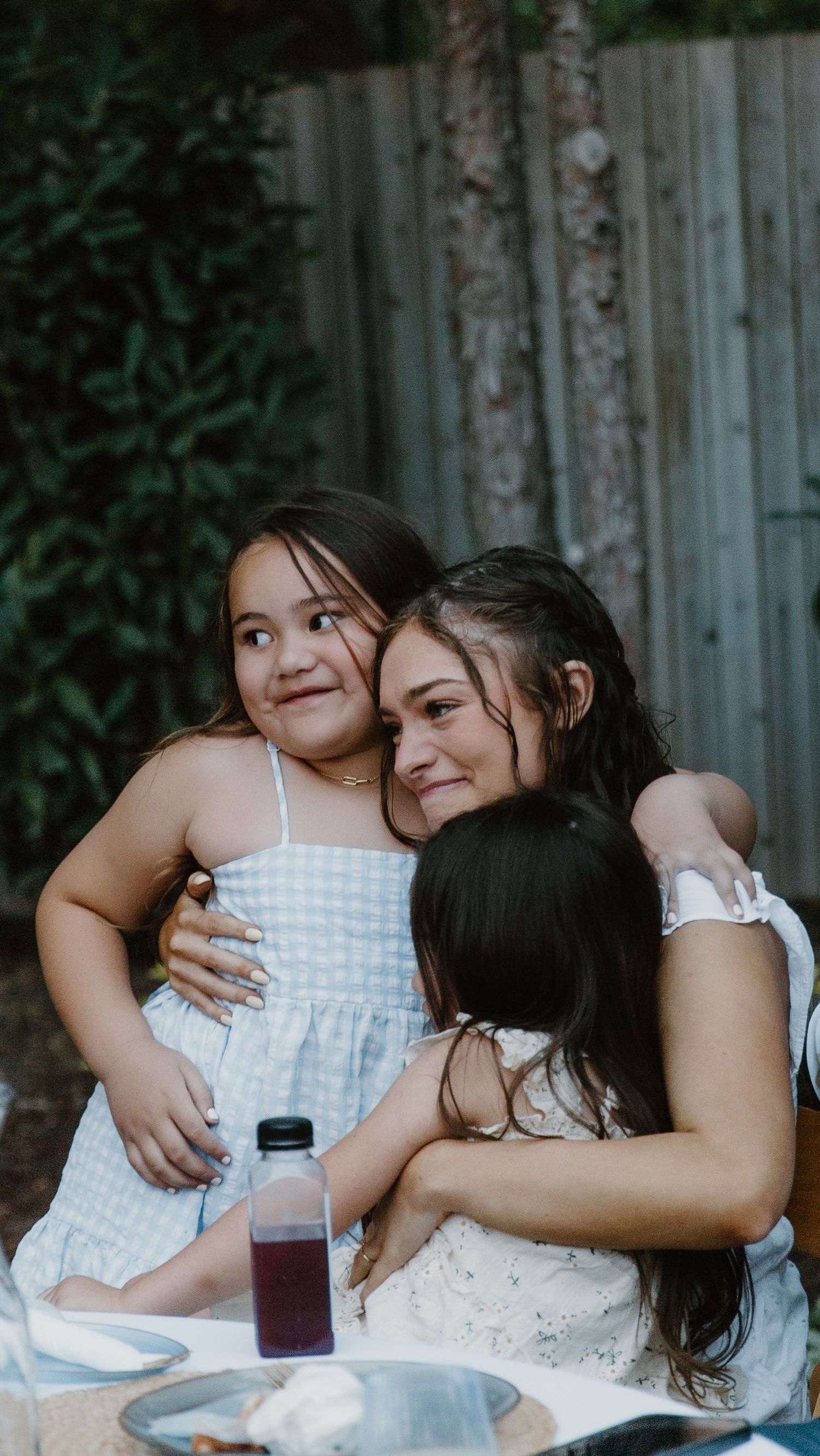 Two young girls in white dresses share affectionate hugs during an outdoor gathering.