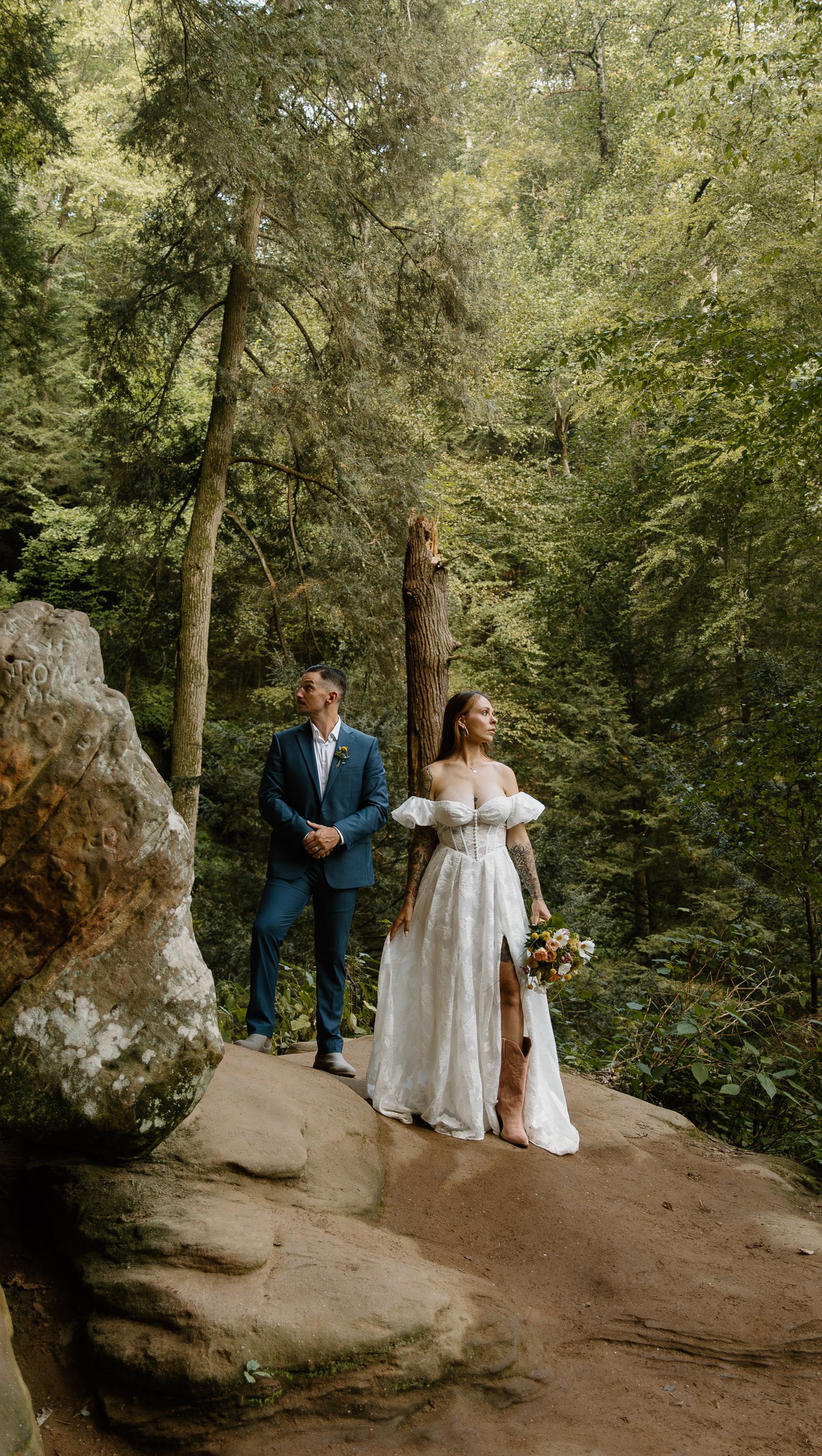 A couple stands together on a large boulder surrounded by lush green forest during their wedding photoshoot.