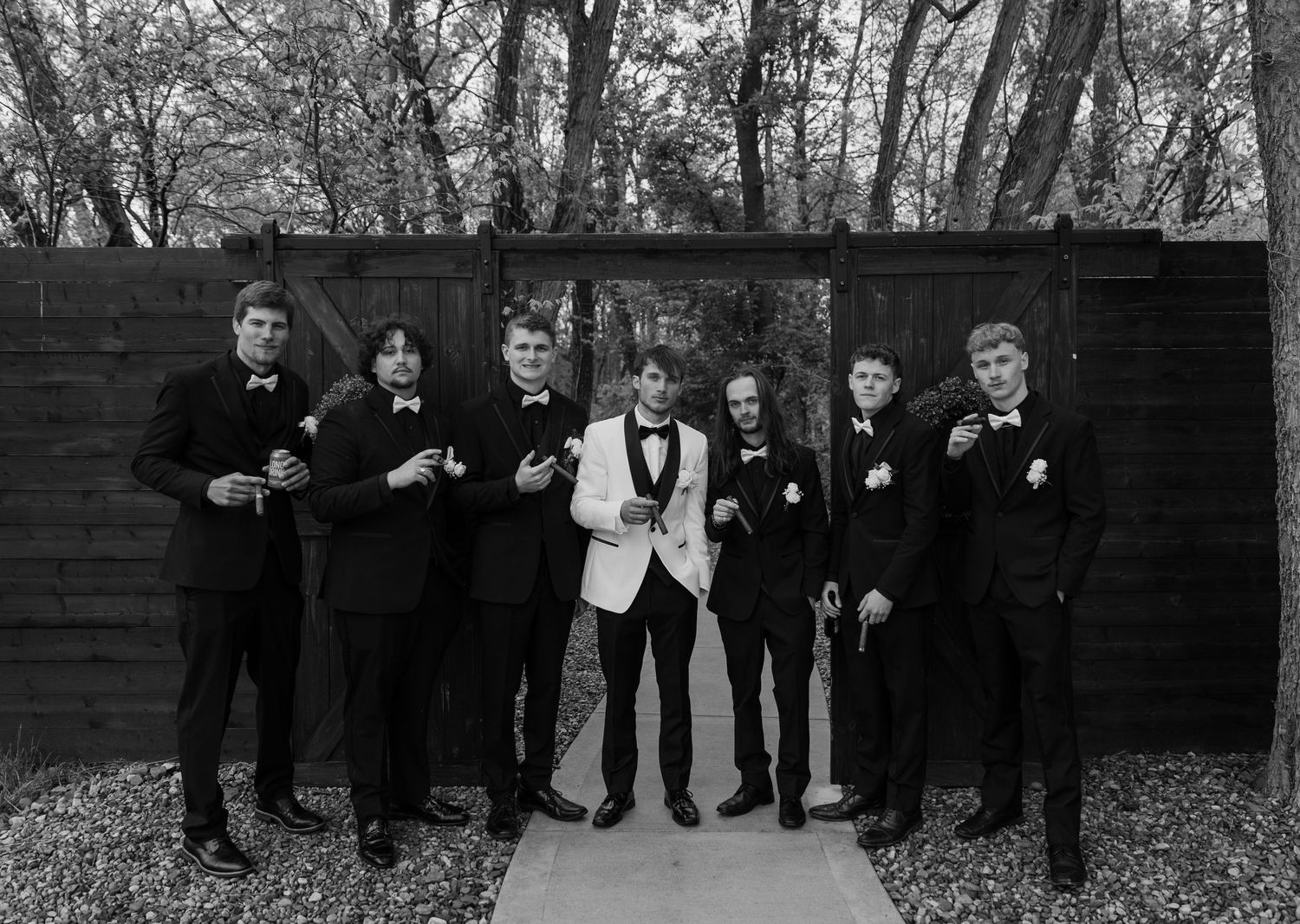 Group of seven young men in black tuxedos with bow ties posing outdoors against dark wooden wall, black and white.