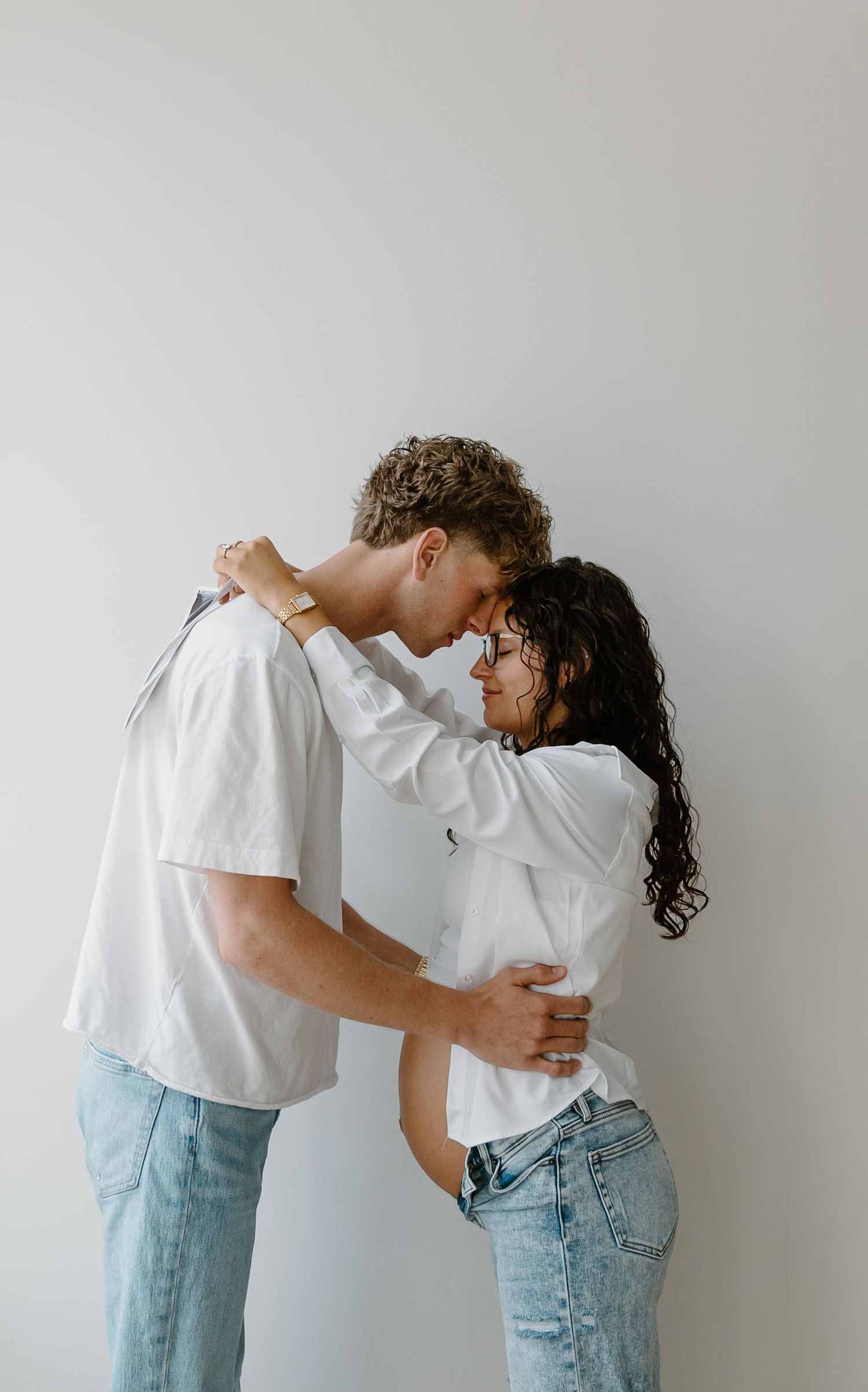 A young couple in white shirts and jeans share a romantic embrace, touching foreheads tenderly against a light gray wall.