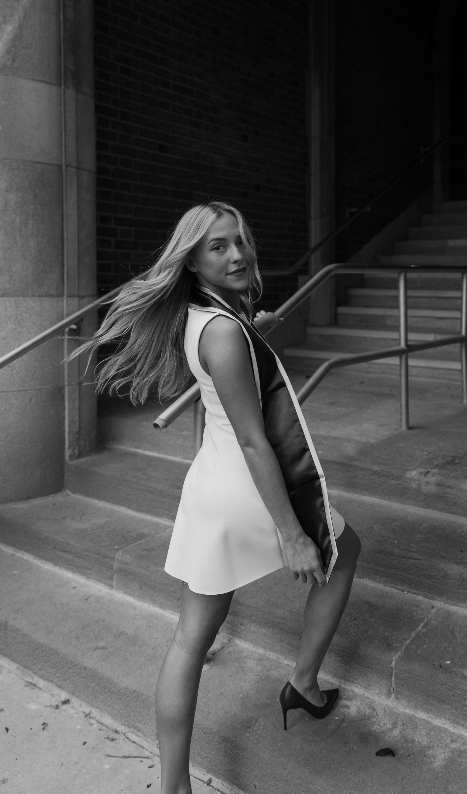 Black and white photo of a young blonde woman in a white dress posing on stone steps outside a brick building.