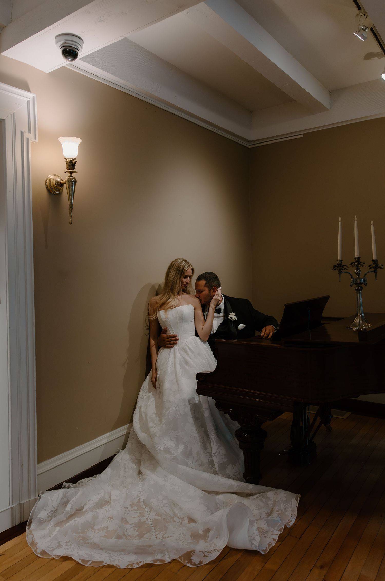 Bride and groom share an intimate moment beside a grand piano in a warmly lit elegant room.