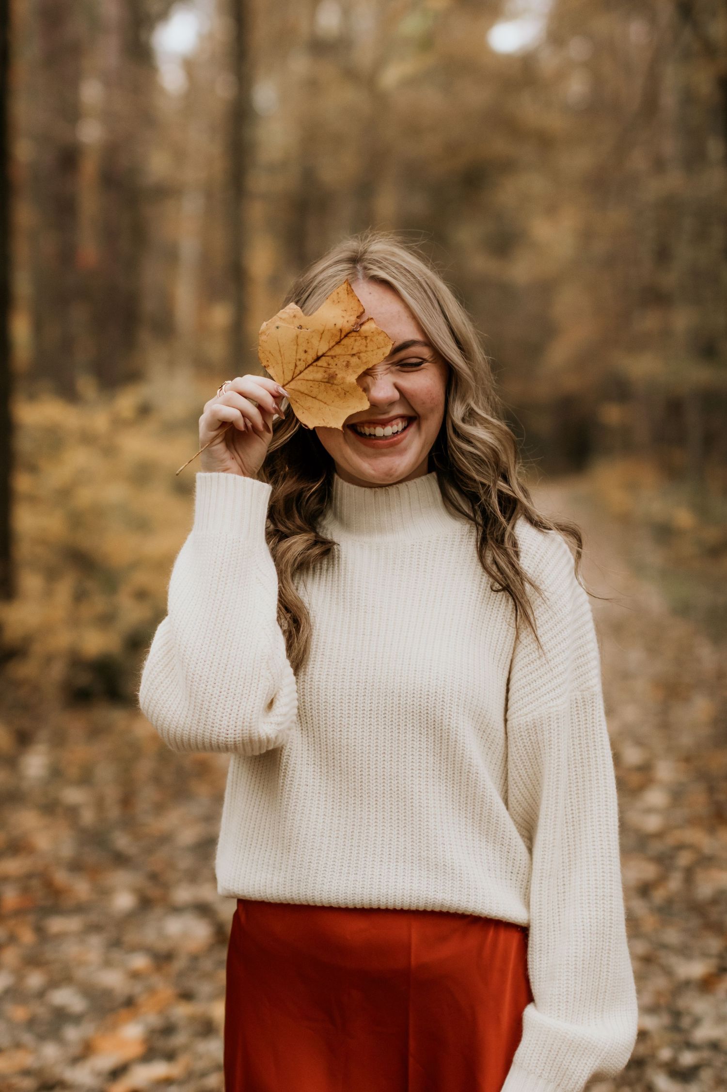 Playful autumn portrait with subject holding a fallen maple leaf while wearing a cream sweater.