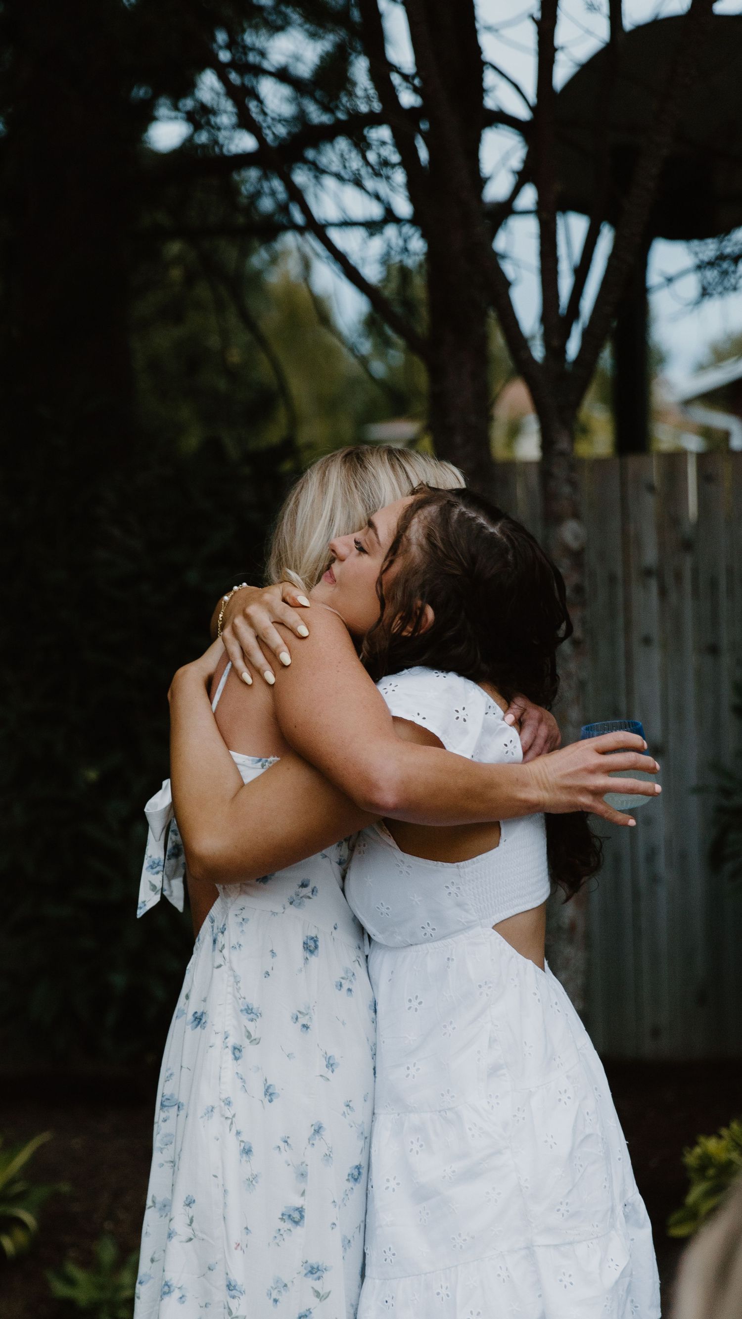 Two people in light-colored dresses sharing an emotional embrace outdoors.