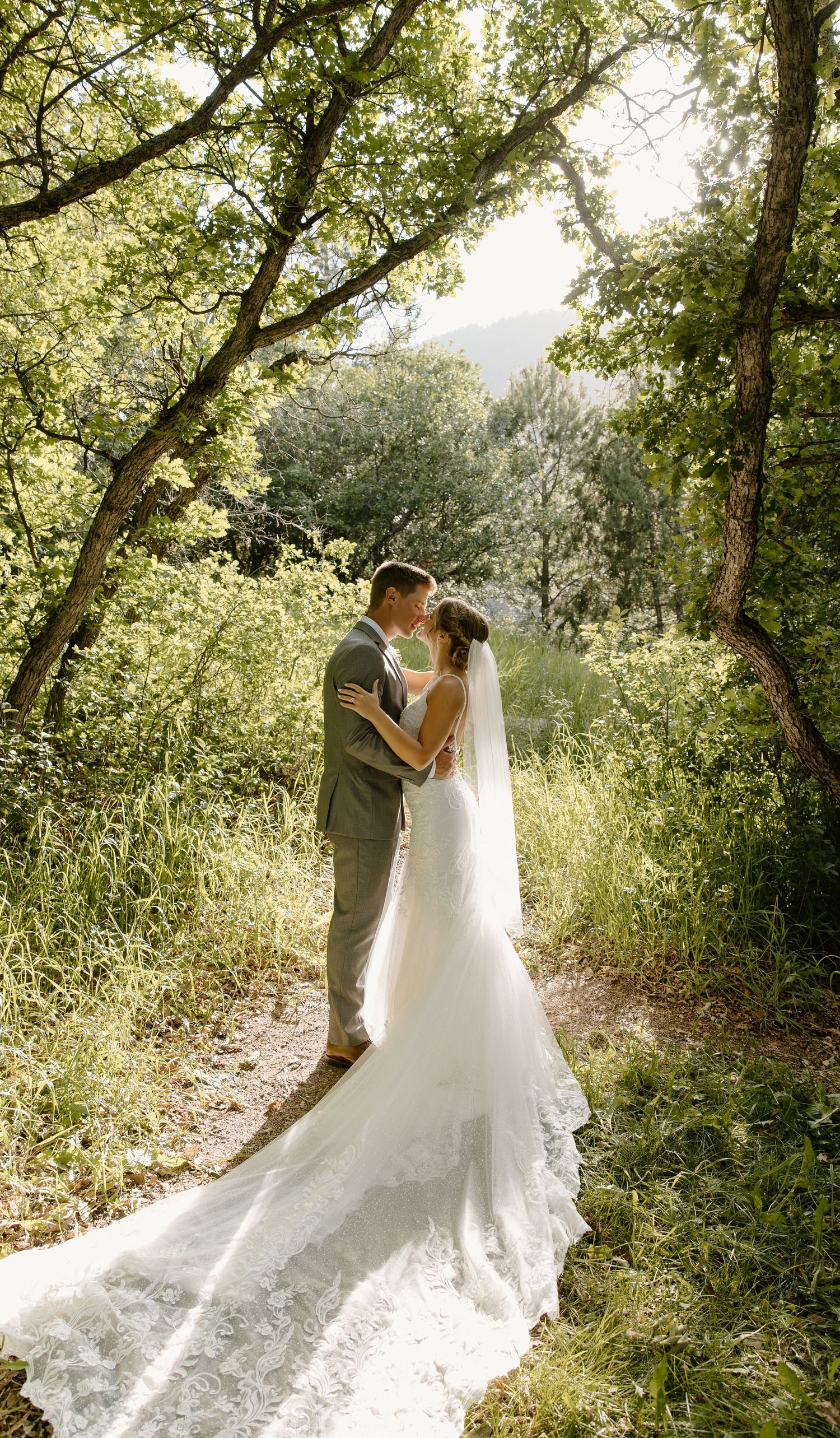 A romantic wedding moment captured in a sunlit forest setting with a flowing white dress trailing on the ground.