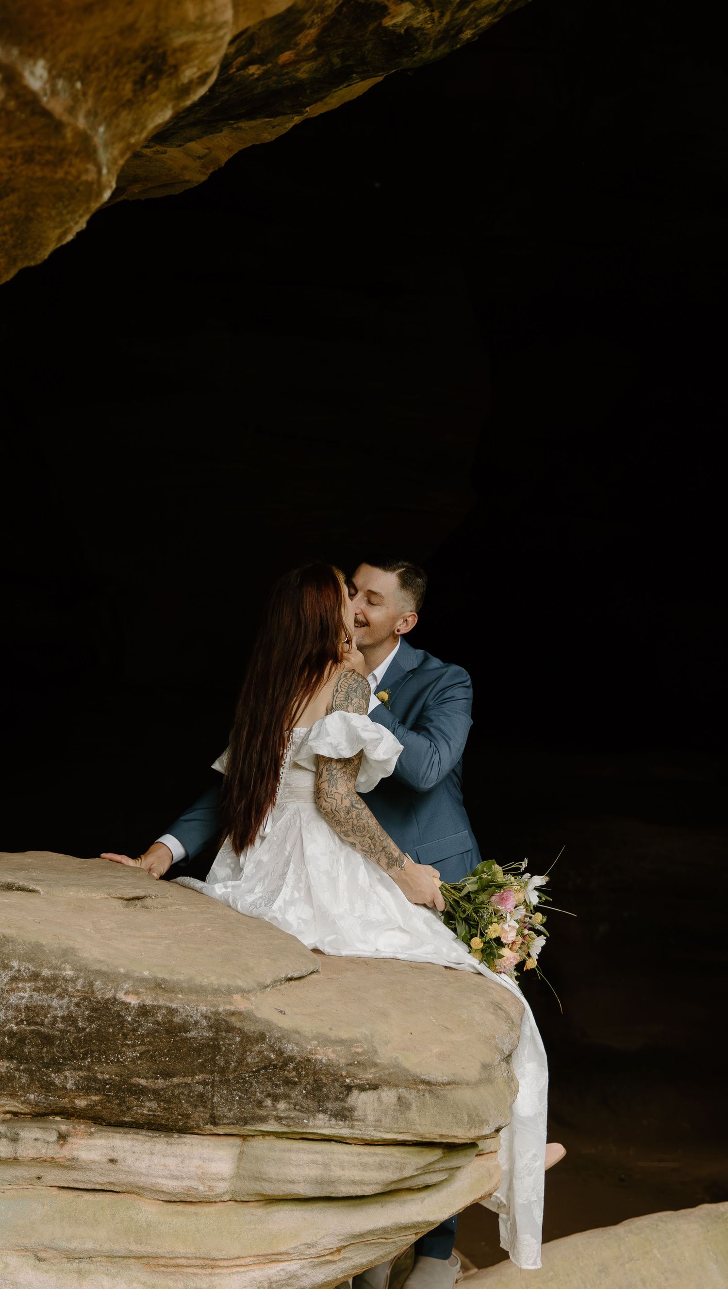 A couple in wedding attire share an intimate moment while sitting on a rock formation inside a dimly lit cave.
