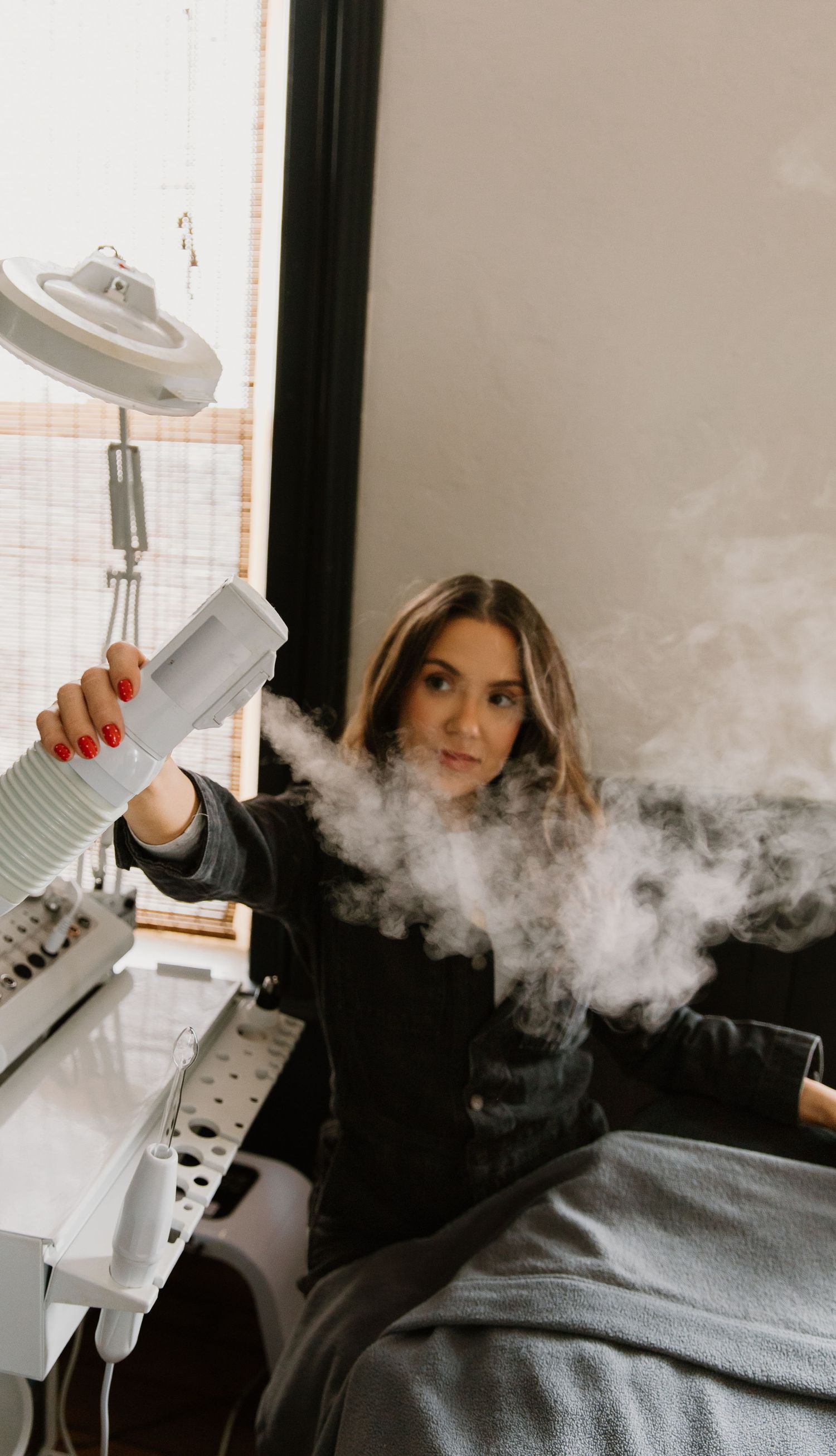 A woman in a black outfit holds a steamer device releasing white steam in a modern beauty salon setting.