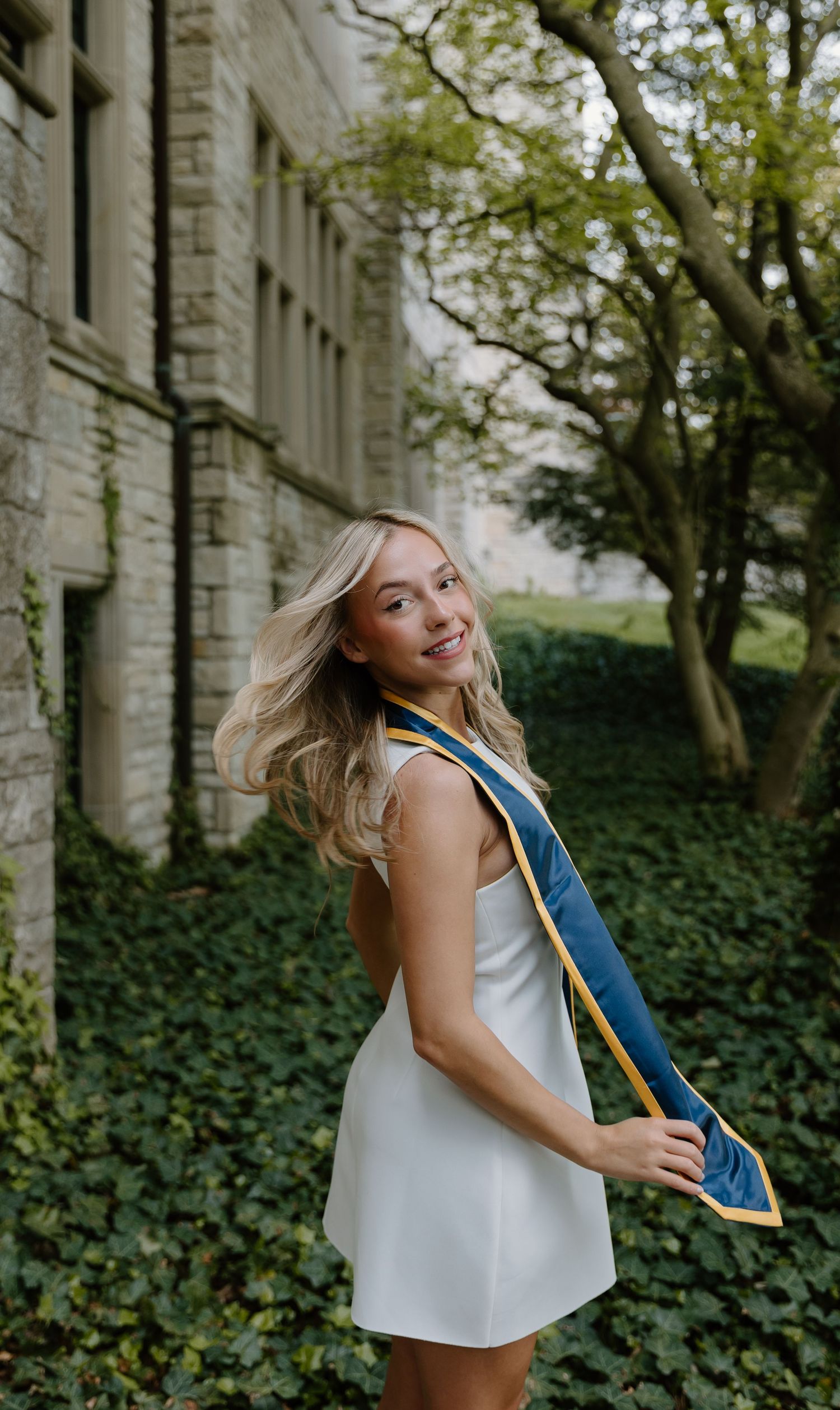 Smiling blonde graduate in white dress with blue and gold sash poses outdoors near stone building and green trees.