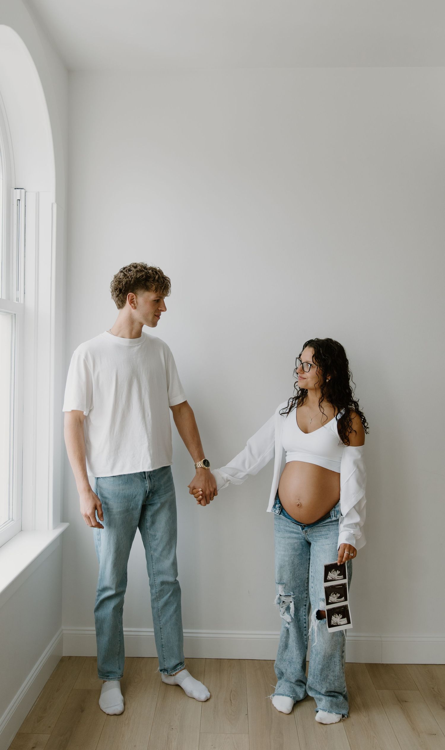 Pregnant woman holding ultrasound photos while holding hands with her partner near a bright window in a white room.