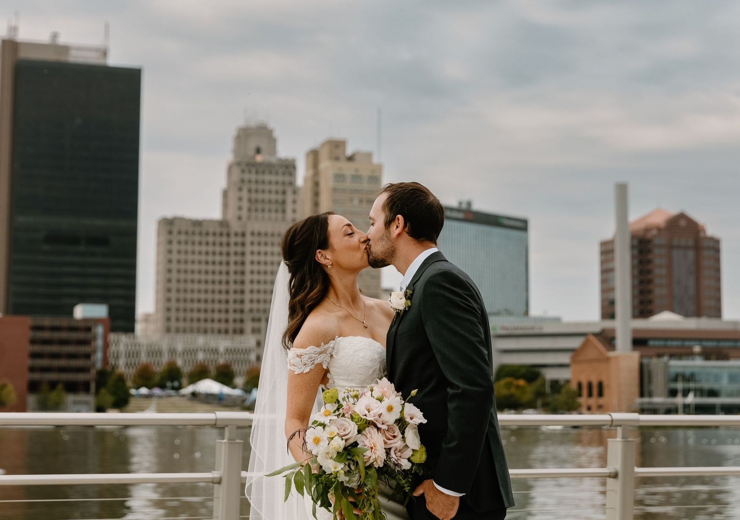 A couple shares a romantic kiss on their wedding day against a cityscape backdrop with tall buildings and water.
