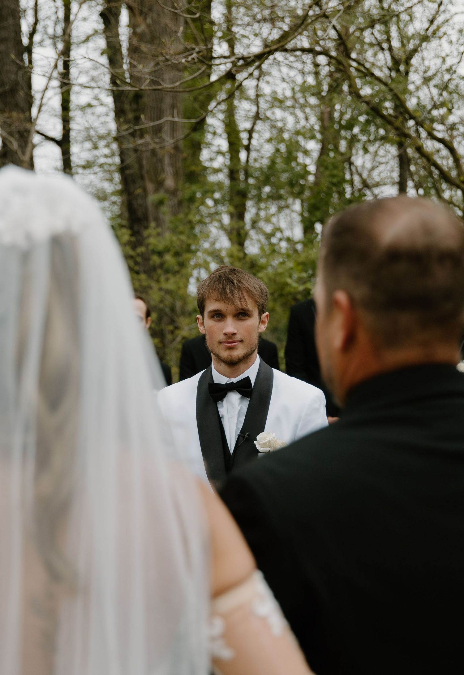 Emotional groom in white tuxedo watches bride approach at outdoor woodland wedding ceremony.