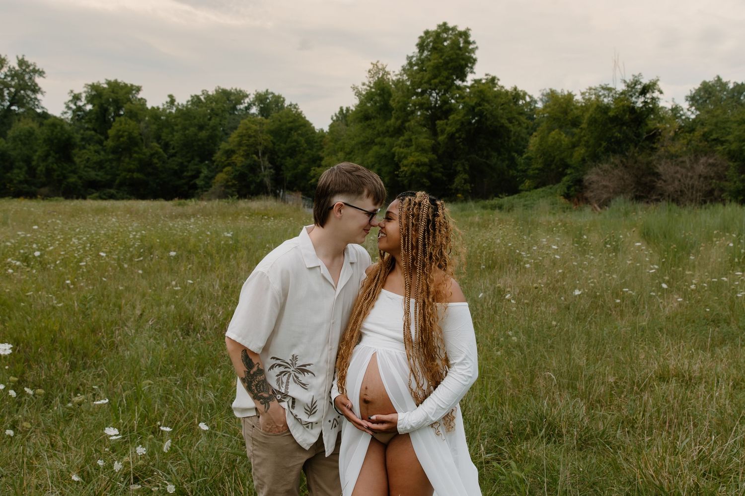 Couple in white outfits sharing a tender moment in a wildflower meadow, showcasing a maternity photoshoot.