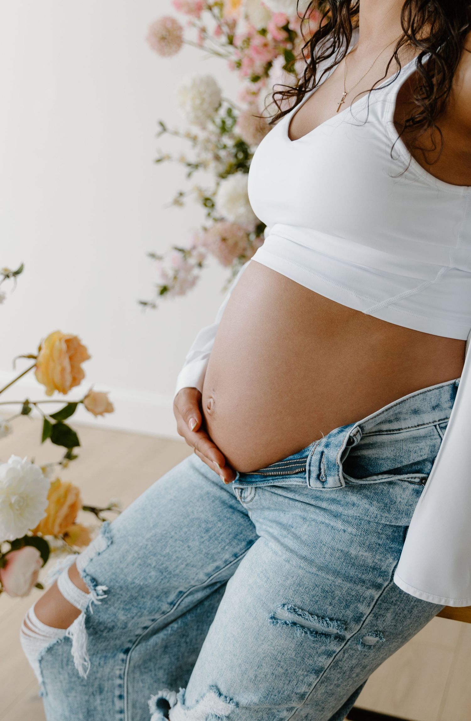 Pregnant woman in white crop top and ripped jeans cradling baby bump, surrounded by soft floral arrangement.